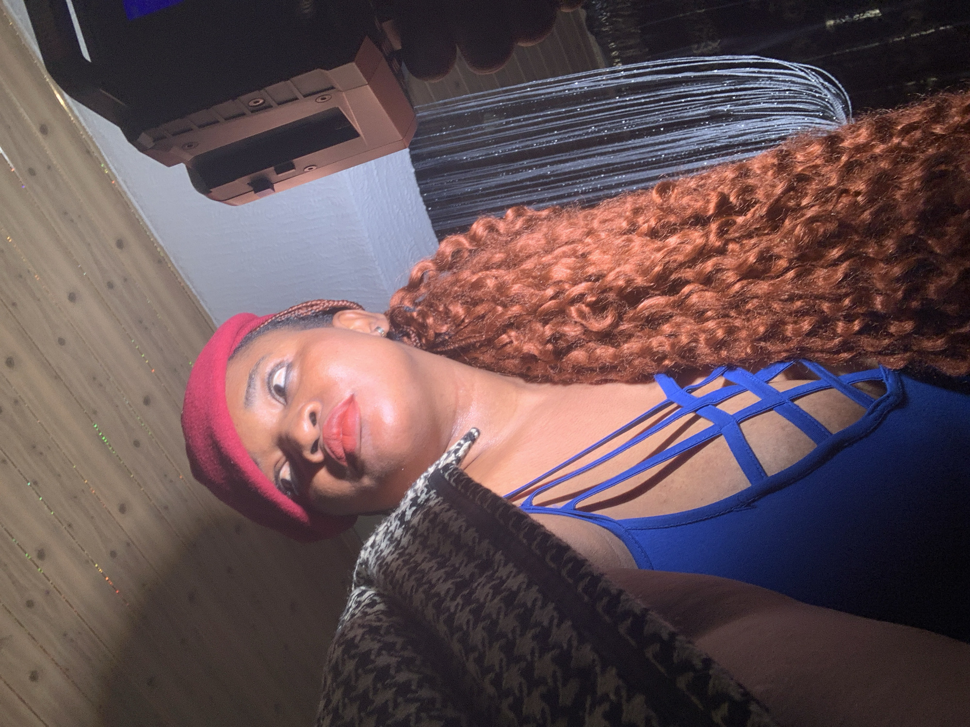 A medium shot captures a person, likely a woman, posing indoors. She has dark skin and is wearing a red beret on her head. Her prominent, long, and voluminous hair is reddish-brown and very curly, flowing down towards her back. She is dressed in a royal blue top featuring an intricate criss-cross strap design across the upper chest. A black and white patterned jacket, possibly houndstooth, is draped over her left shoulder and arm. Her lips are slightly pursed, and her gaze is directed towards the camera, conveying a confident or alluring expression.
The background is varied, suggesting a decorated indoor setting. In the upper left, a dark, bulky electronic device or piece of machinery is visible, possibly mounted on a ceiling or high wall. Behind the person's hair, a dense arrangement of thin, horizontal, reflective lines or strands creates a decorative effect, possibly tinsel or fiber optics. To the left of her face, a wall appears to be constructed of wooden planks, with small, colorful, glittery reflections scattered across its surface. The lighting is artificial, creating strong highlights on her face and hair, and suggesting a dimly lit space, potentially during the evening. The overall scene implies a social environment within Barakin Akawo, Nigeria. No discernible text is visible in the image.