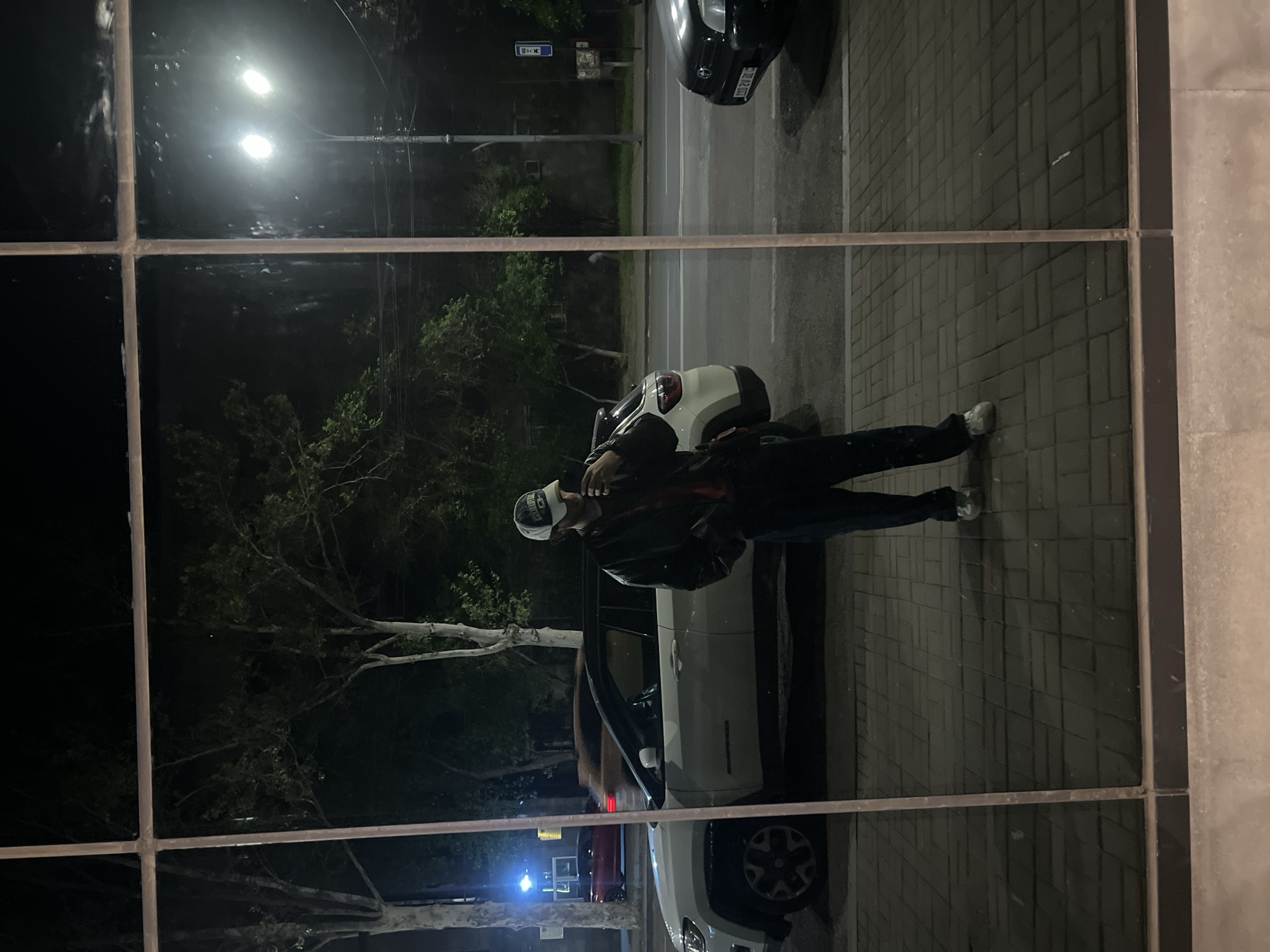 A medium shot shows a man standing on a sidewalk next to a white Jeep, as viewed through a window frame. The man wears a black leather jacket, dark pants, and a white cap. He faces left, looking towards the front of the Jeep with the driver-side door open. Behind the Jeep, a red car is visible. In the background, a street with trees, streetlights, and other cars is seen. The location is likely Yerevan, Armenia.