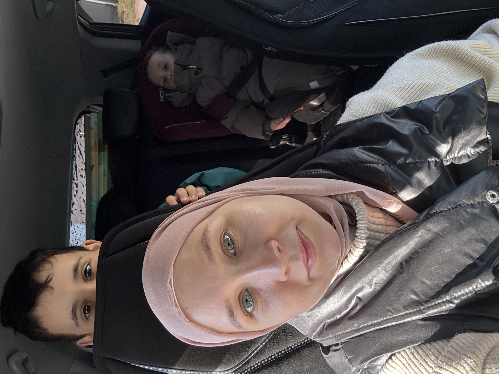 The image is taken inside a car. There is a woman wearing a hijab in the foreground, looking at the camera. Next to her is a young boy, whose face is partially visible. In the back seat, a baby is strapped into a car seat. Other details include a window on the left side and what looks like a black car seat in the background. It is impossible to ascertain the location as the city of Chaikovsky, Russia.