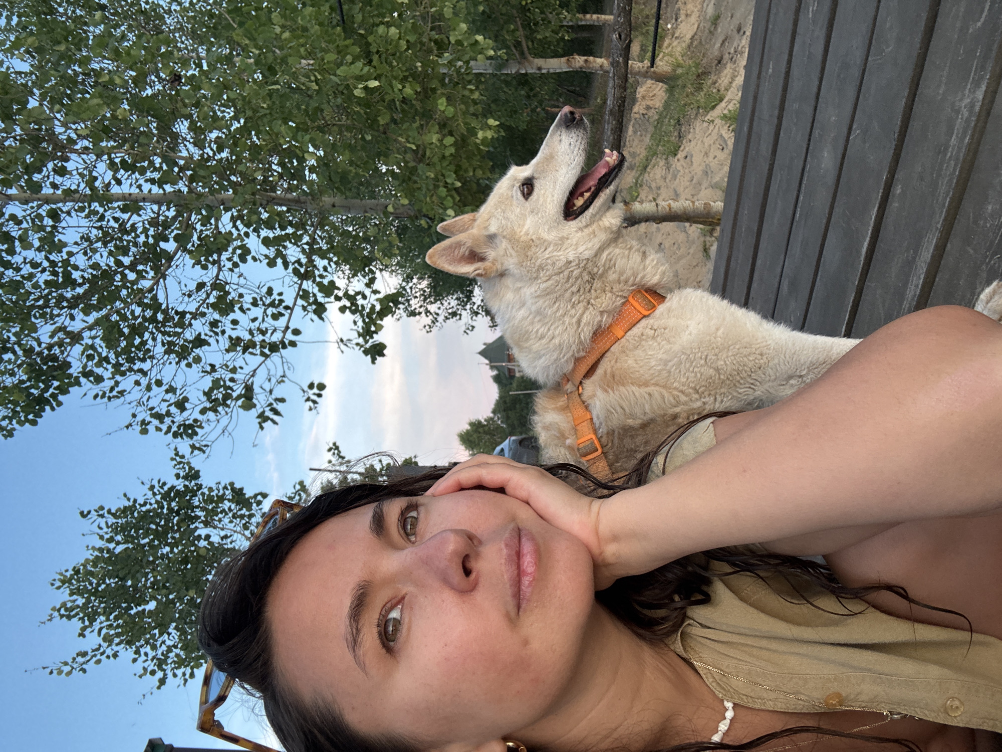 A medium shot captures a woman and a dog sitting on a wooden bench outdoors. The woman is in the foreground on the left, leaning her head on her hand and looking to the left. She has long, dark, wavy hair and is wearing a tan button-up shirt and a white beaded necklace. Her right arm is resting on the dog. The dog, a light-colored husky mix, is sitting next to her, facing to the right with its mouth open. It is wearing an orange harness. The background shows trees and a sandy area, consistent with a park or natural setting. The sky is blue with some clouds. The image appears to have been taken in the city of Kamysh, Russia.