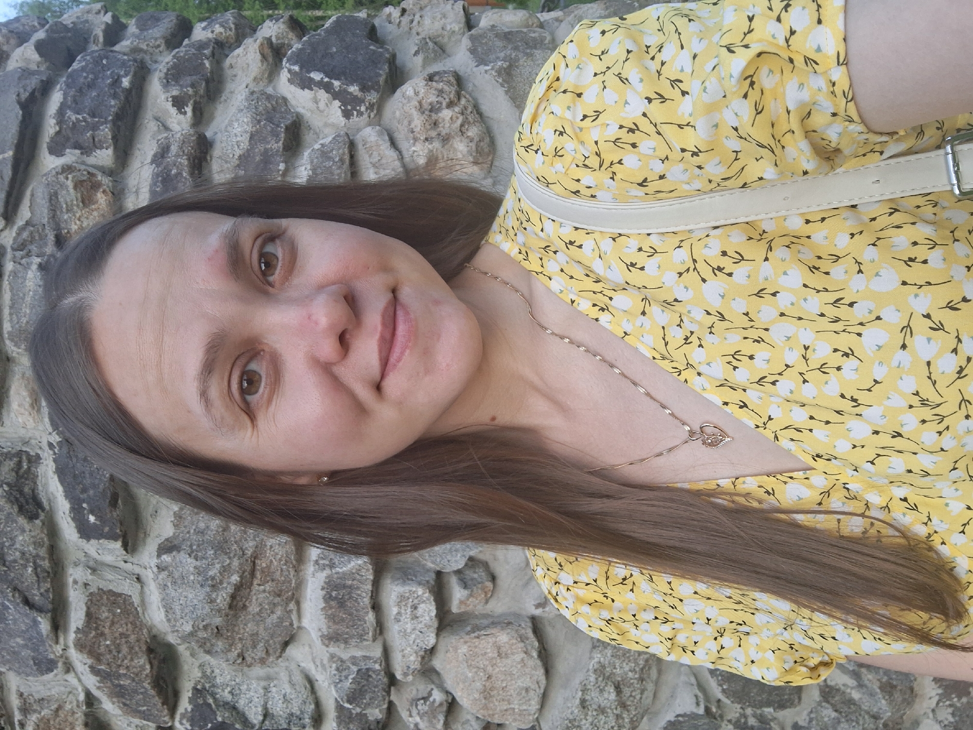 A woman with brown hair and fair skin is posing for a photo in front of a stone wall. She is wearing a yellow floral dress and a delicate gold necklace with a pendant. A strap, possibly from a bag, is visible over her shoulder. The background appears to be a stone wall consisting of various sized rocks. The location is Chaikovsky, Russia.