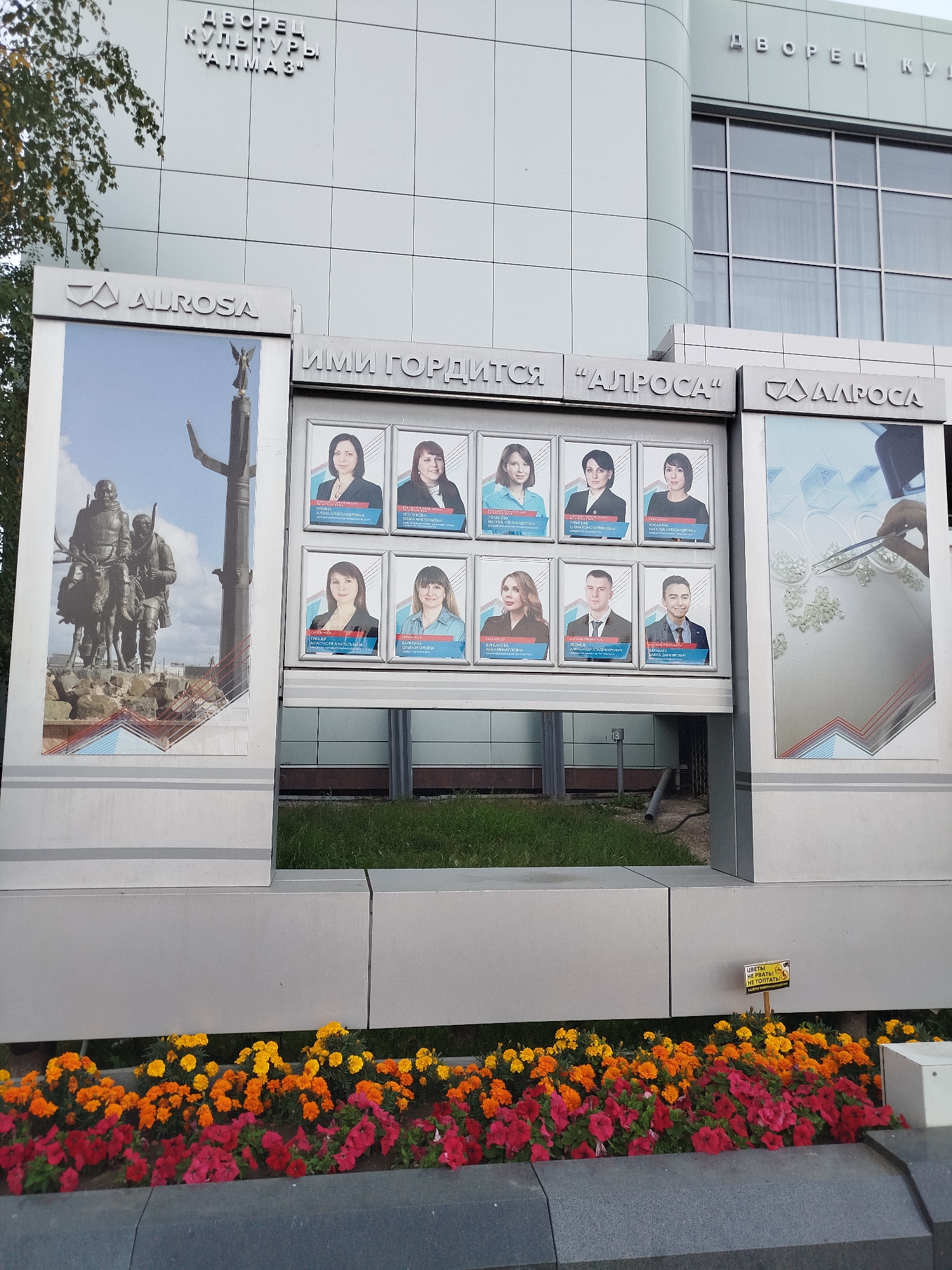 The image shows the exterior of the "Almaz" Palace of Culture in Mirny, Russia, with a promotional display for ALROSA, a diamond mining company. The display features a large image of a statue of two men on a reindeer-drawn sleigh, and a grid of portraits of employees with their names and titles. There are also images related to diamond cutting. The display is surrounded by flower beds with red, orange, and yellow flowers. The building is modern and appears to be made of concrete and glass. The sky is overcast.