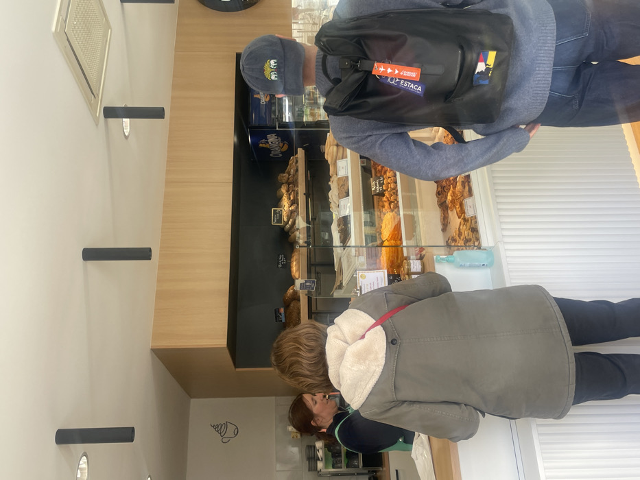 The scene depicts the interior of a bakery or patisserie located in Montigny-le-Bretonneux, France. Three individuals are present: one female employee behind a light wood counter, a female customer facing the counter, and a male customer positioned to the right of the female customer, also facing the counter with his back to the camera.
The female employee, with reddish-brown hair, wears a dark top and a light green apron, engaging with the female customer. The female customer wears a khaki-colored long jacket with a cream-colored fleece lining visible at the collar and a red strap across her shoulder, over dark pants. She stands directly in front of the counter.
To the right, the male customer wears a grey long-sleeved shirt, dark blue jeans, and a navy blue baseball cap with a yellow and green multi-elephant logo on the front. He carries a black backpack with two keychains attached; one is blue with white text "POUR ESTACA" and red airplane graphics, the other is red with white text "REMOVE BEFORE FLIGHT".
Behind the counter and customers, glass display cases showcase various baked goods, including breads, pastries, and what appear to be individual desserts or tarts. A blue bottle of hand sanitizer is visible on the counter. Above the display cases, shelves hold additional bread items, and a refrigerator with an "Orangina" logo is visible. The walls are light wood paneling above dark shelving, with a stylized coffee cup silhouette decal on the left wall. The ceiling is white, featuring multiple dark cylindrical track lights and a rectangular air conditioning vent.