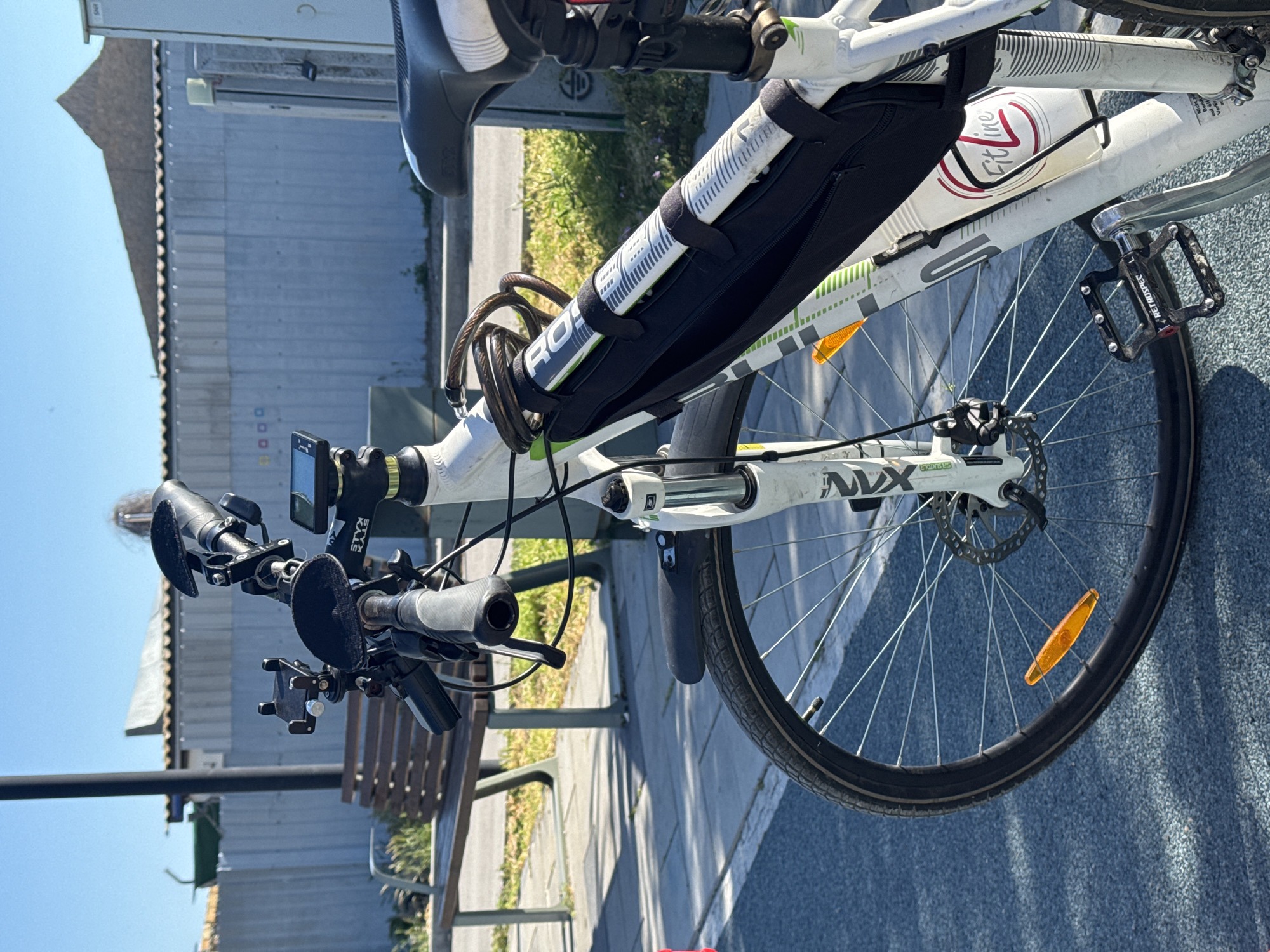 The image shows a close-up of a white bicycle parked outdoors, with a bench visible in the background. The bike has a black frame bag, a water bottle cage with a red and white bottle, and accessories on the handlebars, including a digital display and a phone mount. The front wheel has a disc brake and an orange reflector. The bicycle pedal is from the brand "RETROSPEC". The background includes a building and some greenery. The location is a paved area with a bench, potentially within the city limits of Городское поселение Щелкино, Ukraine. No people are visible.
