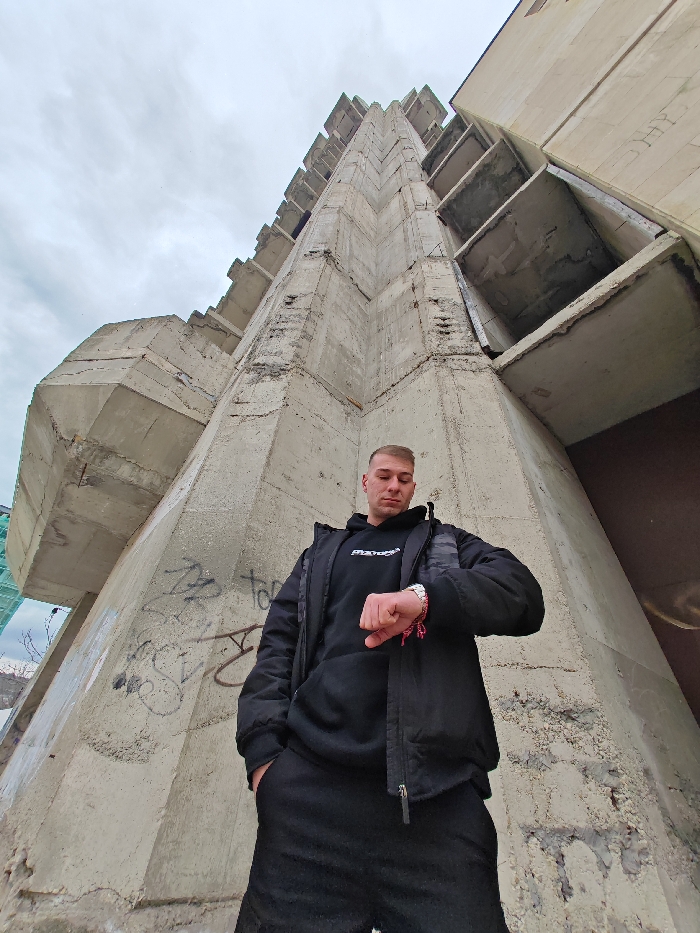 A single adult male is depicted standing in front of a monumental concrete structure, identified as the Monument to 1300 Years of Bulgaria in Shumen, Bulgaria. The man, with short, light-colored hair, is positioned centrally in the lower frame, facing slightly left of the camera and looking downwards at his left wrist. He is wearing a black hooded sweatshirt with a white logo, a black unzipped puffer jacket, and dark trousers, with his right hand in his pocket. A metallic wrist watch and a multi-strand red/pink bracelet are visible on his left wrist. The background consists of a massive, angular, Brutalist-style concrete pylon of the monument, extending diagonally upwards and to the right, showing exposed aggregate, spalling, and graffiti tags in black and white paint. The upper portion of the image is dominated by an overcast, cloudy sky. A light green structural element, possibly scaffolding, is partially visible on the far left edge of the frame.