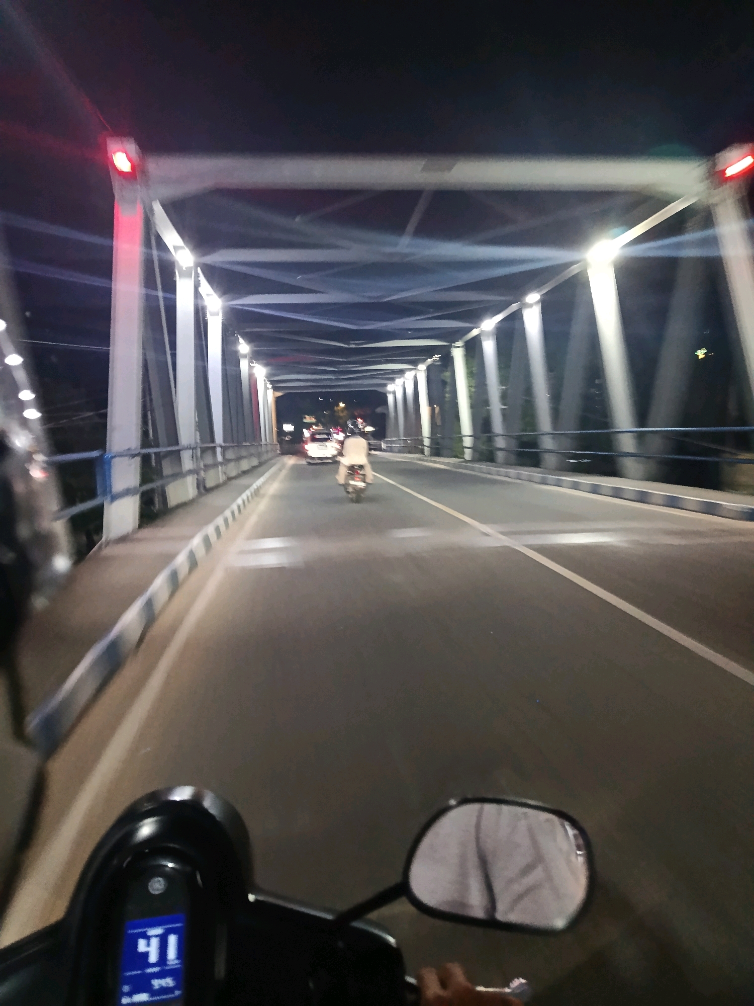 The image depicts a nighttime view from the perspective of someone riding a scooter or motorcycle across a bridge. The bridge is illuminated by a series of lights attached to its structure, which is made of white beams forming a lattice overhead. There are red lights visible on the top corners of the bridge structure. A blue and white striped railing runs along the side of the bridge, separating the roadway from the pedestrian walkway.
On the roadway, another individual is riding a scooter or motorcycle ahead, wearing light-colored clothing. In the distance, a white car is also visible. The road is asphalt and has a white line marking the lane. The scene appears to be within the city of Arjowilangun, Indonesia, based on the provided context. The rider's scooter dashboard is visible in the foreground, displaying the number "41". A rearview mirror reflects the rider's light-colored shirt. The overall atmosphere is that of a well-lit bridge at night, with ongoing traffic.