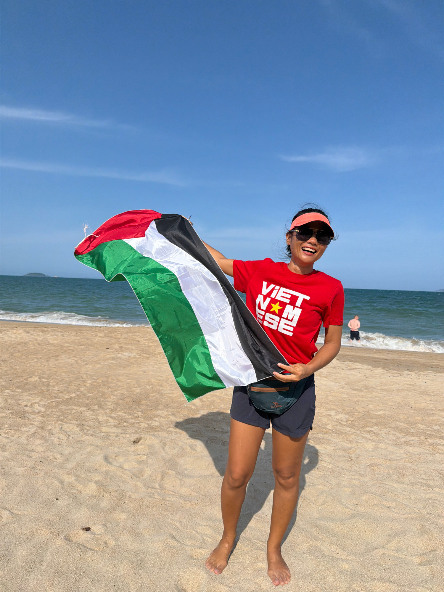 A Vietnamese woman stands on a sandy beach in Nha Trang, Vietnam, holding a Palestinian flag. She is wearing a red t-shirt with "VIETNAMESE" printed in white, a black fanny pack, and dark shorts. She is also wearing sunglasses and an orange visor. The ocean is visible in the background with waves breaking on the shore. The sky is clear and blue with some wispy clouds. A man is standing in the water in the background. The image was taken on a sunny day.