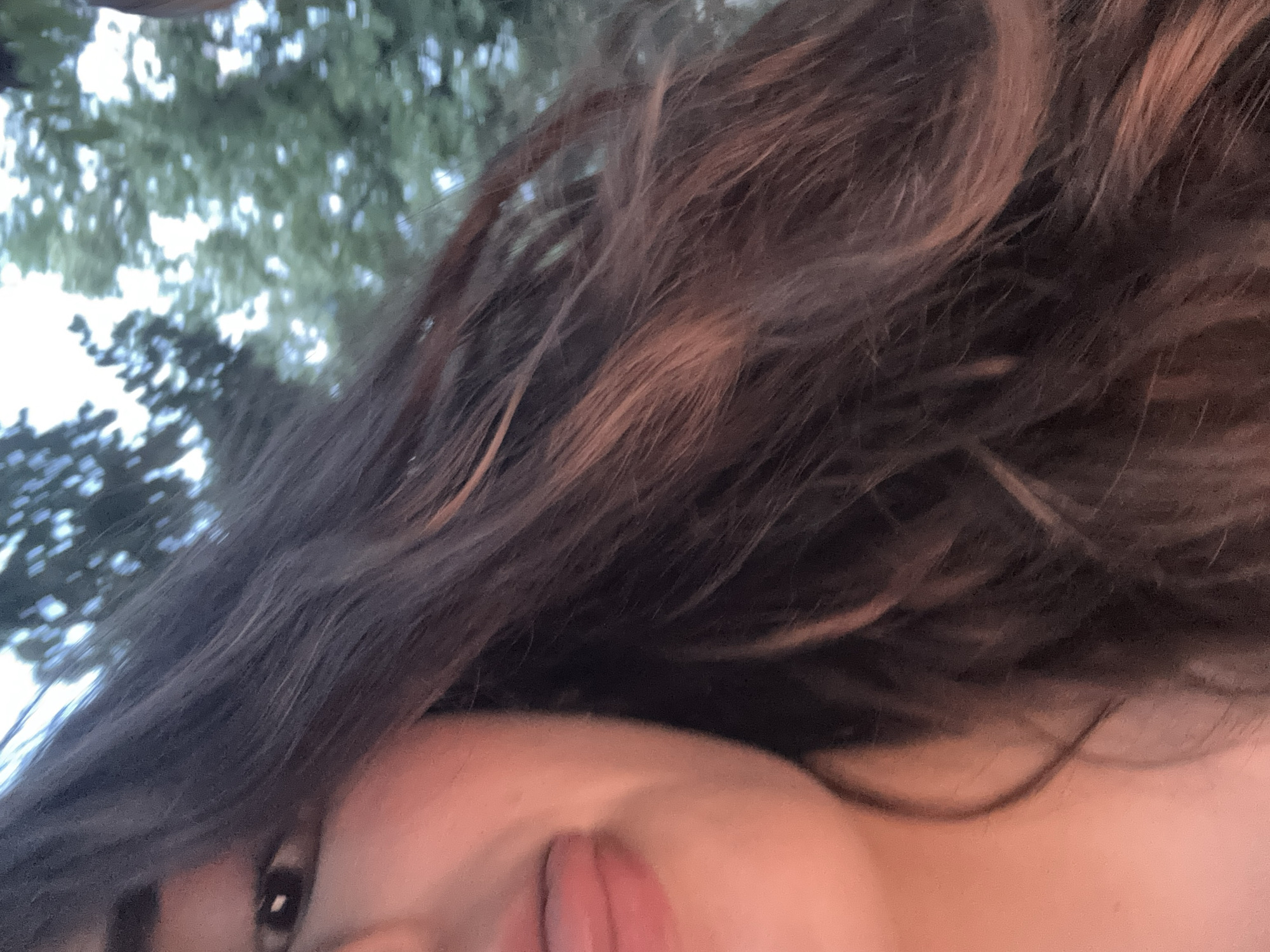 A close-up, cropped image shows a portion of a person's face and hair. The person's left eye is visible, looking towards the viewer. Their mouth is open with their tongue sticking out. Dark brown hair, with reddish highlights, fills the majority of the frame, obscuring the background. The background appears to be trees and foliage, suggesting an outdoor setting. The image is not specific to Shumen, Bulgaria.