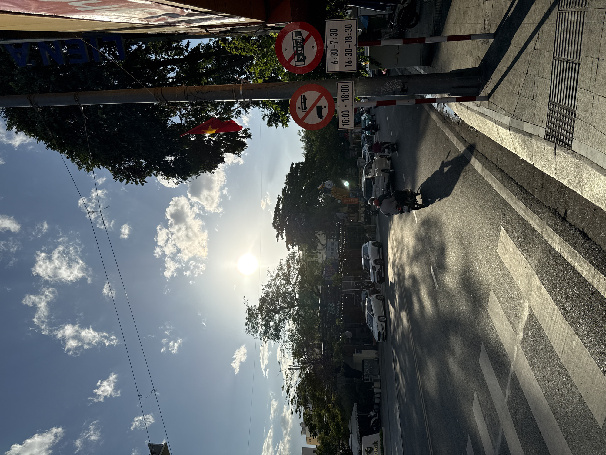 The image depicts a street scene in Nha Trang, Vietnam, bathed in the warm light of a setting or rising sun. The sun, a bright orb, is positioned in the upper left quadrant of the sky, casting long shadows across the asphalt road. A pedestrian crossing is visible in the foreground, with white stripes marking its path.
On the road, several vehicles are present, including cars and motorcycles. A few individuals are riding motorcycles, their forms silhouetted against the light. In the background, a row of buildings with awnings and trees line the street. A red Vietnamese flag with a yellow star flies from a pole.
Traffic signs are prominently displayed on a pole on the right side of the image. One sign is circular with a red border and a diagonal red line, indicating a prohibition. Inside, it shows a silhouette of a bus and a truck, suggesting a restriction on these types of vehicles. Another circular sign has similar markings but also contains the text "> 29 CHO", which translates to "over 29 seats" or "over 29 passengers", further specifying the restriction for buses. Below these signs, rectangular plates indicate specific time windows for these restrictions: "16:00 - 18:00" and "6:30 - 7:30", "16:30 - 18:30". These signs collectively indicate a timed restriction on certain types of buses or vehicles exceeding a certain passenger capacity.
The environment appears to be an urban setting, with streetlights, power lines, and sidewalks contributing to the cityscape. The presence of pedestrians and vehicles suggests an active street. The overall atmosphere is one of a typical day in a Vietnamese city, captured during the golden hour.