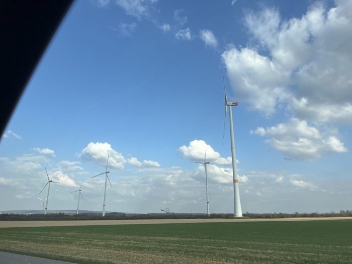 The image displays an expansive rural landscape situated in the vicinity of Eschweiler, Germany, under a bright blue sky with scattered white cumulus clouds. In the foreground, a dark surface, possibly a road or shoulder, occupies the bottom-left, adjacent to a strip of brownish soil that transitions into a large, verdant green agricultural field filling the lower half of the frame.
Multiple wind turbines are prominent subjects within the scene. A tall, white/light gray wind turbine with an orange band around its base and three white blades with red tips stands in the right midground, appearing static. To the left and receding into the distance, at least five additional wind turbines of similar design are visible across the midground horizon, also appearing stationary. A linear band of dark green/brown trees or bushes forms a natural separation between the cultivated fields and the distant background. In the far background, slightly right of the frame's center, large construction equipment, specifically a crane, is discernible against the horizon. No individuals are present within the frame. The upper-left corner of the image is partially obscured by a dark, curved object, consistent with an interior component of a vehicle, indicating the photograph was taken from inside a vehicle.