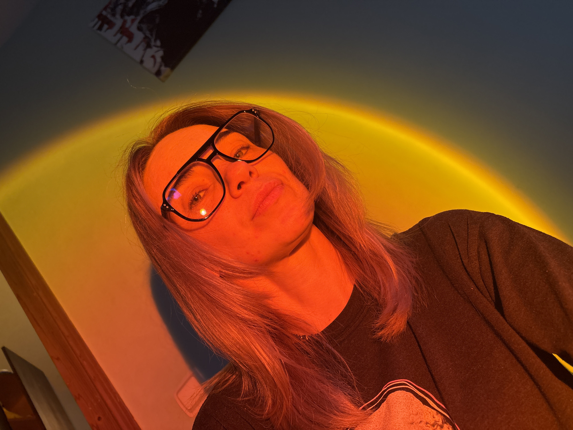 An indoor, eye-level portrait features an individual with long, light-colored hair and black-rimmed eyeglasses, looking up and slightly to the viewer's left with a neutral expression. The person's face and upper torso are illuminated by a strong, warm light source that casts a semi-circular gradient of yellow to orange-red light onto the wall behind them, suggesting the use of a "sunset lamp."
The intense warm lighting bathes the person's skin in a reddish hue and highlights streaks in their hair. They are wearing a dark, long-sleeved top. To the far left, a wooden beam or frame is partially visible, while in the upper left background, a dark, indistinct object, possibly a framed picture or poster, hangs on a dark wall. The ambient light in the upper portion of the image is dim. No visible text is present.