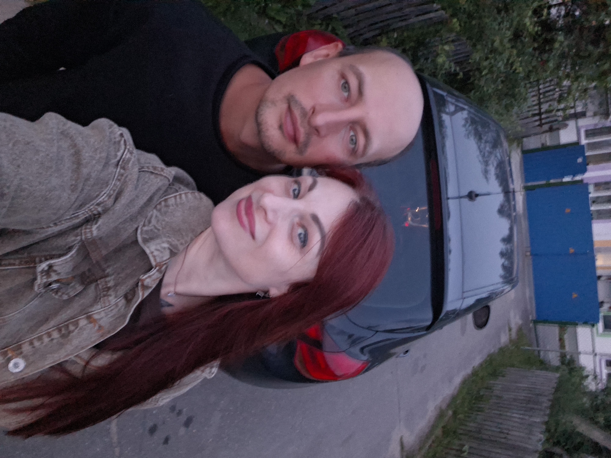 A bald man and a woman with long red hair are taking a selfie outdoors. The man, on the left, is wearing a black shirt and has a light beard. The woman, on the right, is wearing a denim jacket and a necklace. They are standing in front of a dark-colored car. In the background, there is a wooden fence, green bushes, a paved road, and a building with blue doors. The scene appears to be in a residential area, likely within the Забалоцкі сельскі Савет in Belarus, given the context of the query. The lighting suggests it is either early morning or late evening.