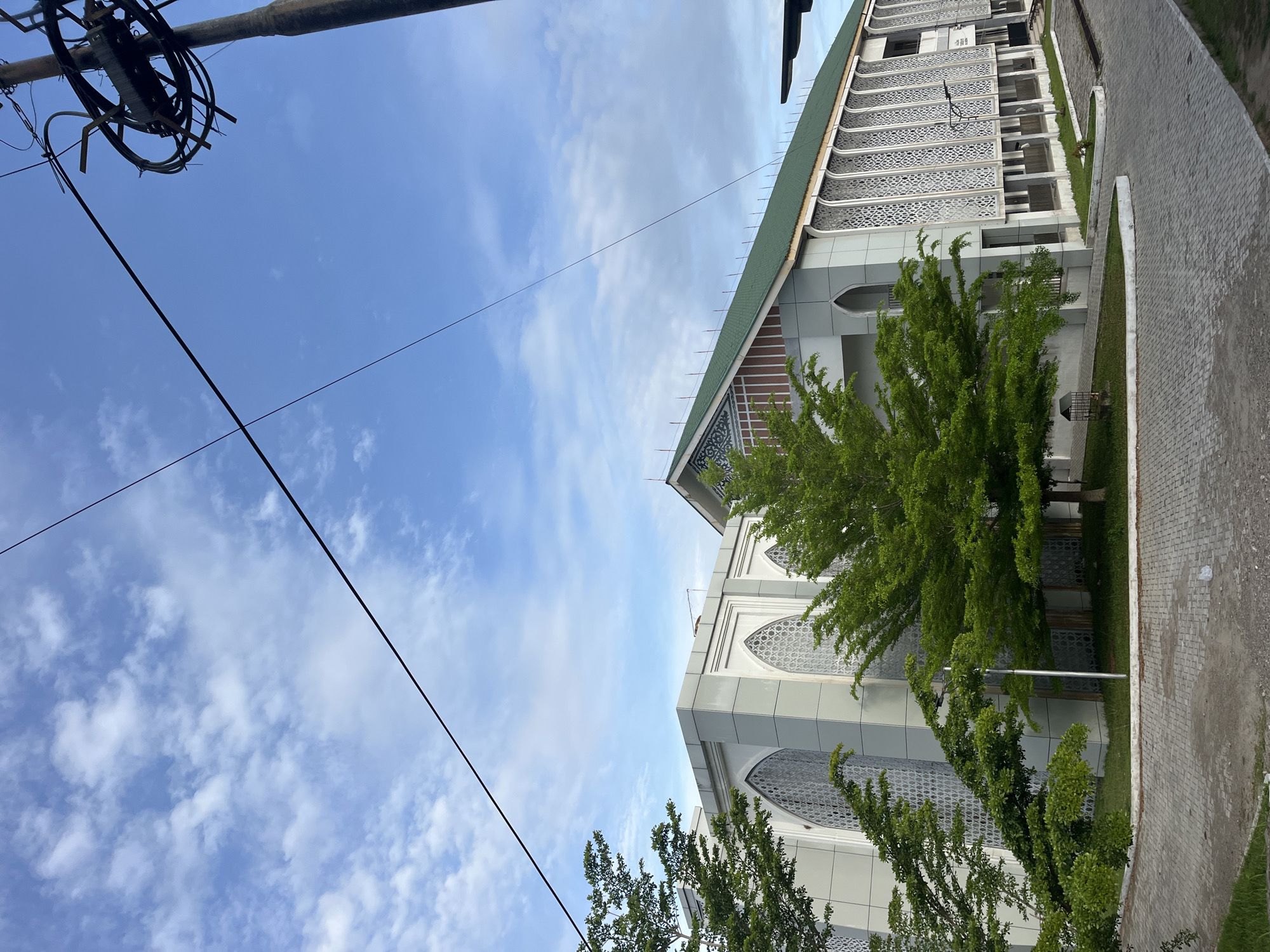The image depicts a building with white and gray architectural features, possibly a mosque, under a partly cloudy sky. Trees with green foliage are visible in front of the building. Overhead power lines cross the upper left of the frame. The image appears to be taken from ground level, looking up at the building. Given the context of Lhokseumawe, Indonesia, it is plausible that this is a local mosque or a similar public building with Islamic architectural elements.