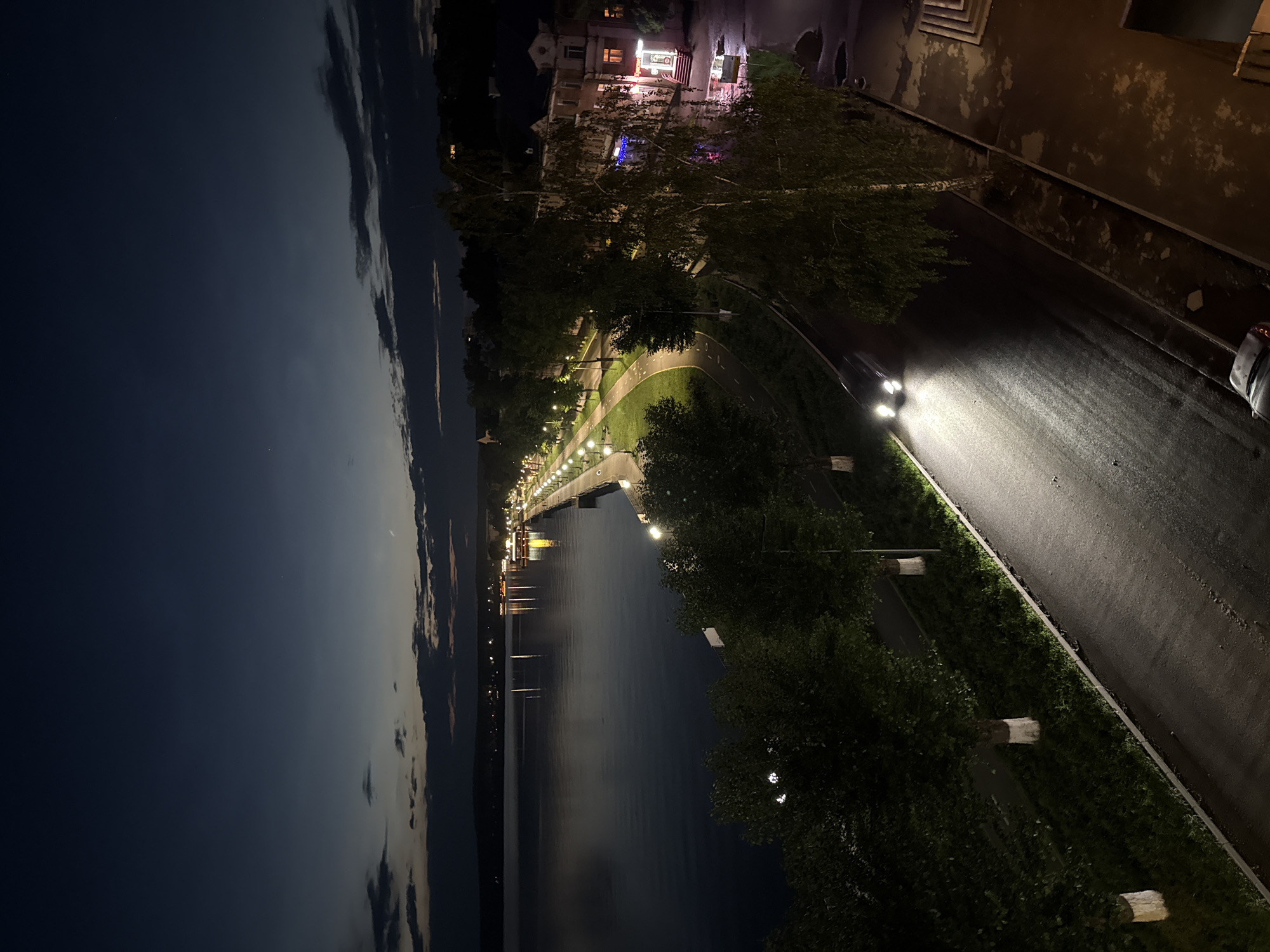 An elevated nighttime, or twilight, view looks down on a wide, calm body of water to the left, bordered by a tree-lined promenade and a road with streetlights. On the right, a two-story building with illuminated windows and balconies is visible. A car with its headlights on drives away from the viewer on the wet asphalt road. The sky is a deep blue, with a few scattered clouds reflecting the light from the horizon. The scene appears to be located along the embankment of Lake Votkinsk in the city of Votkinsk, Russia, known for its scenic waterfront.