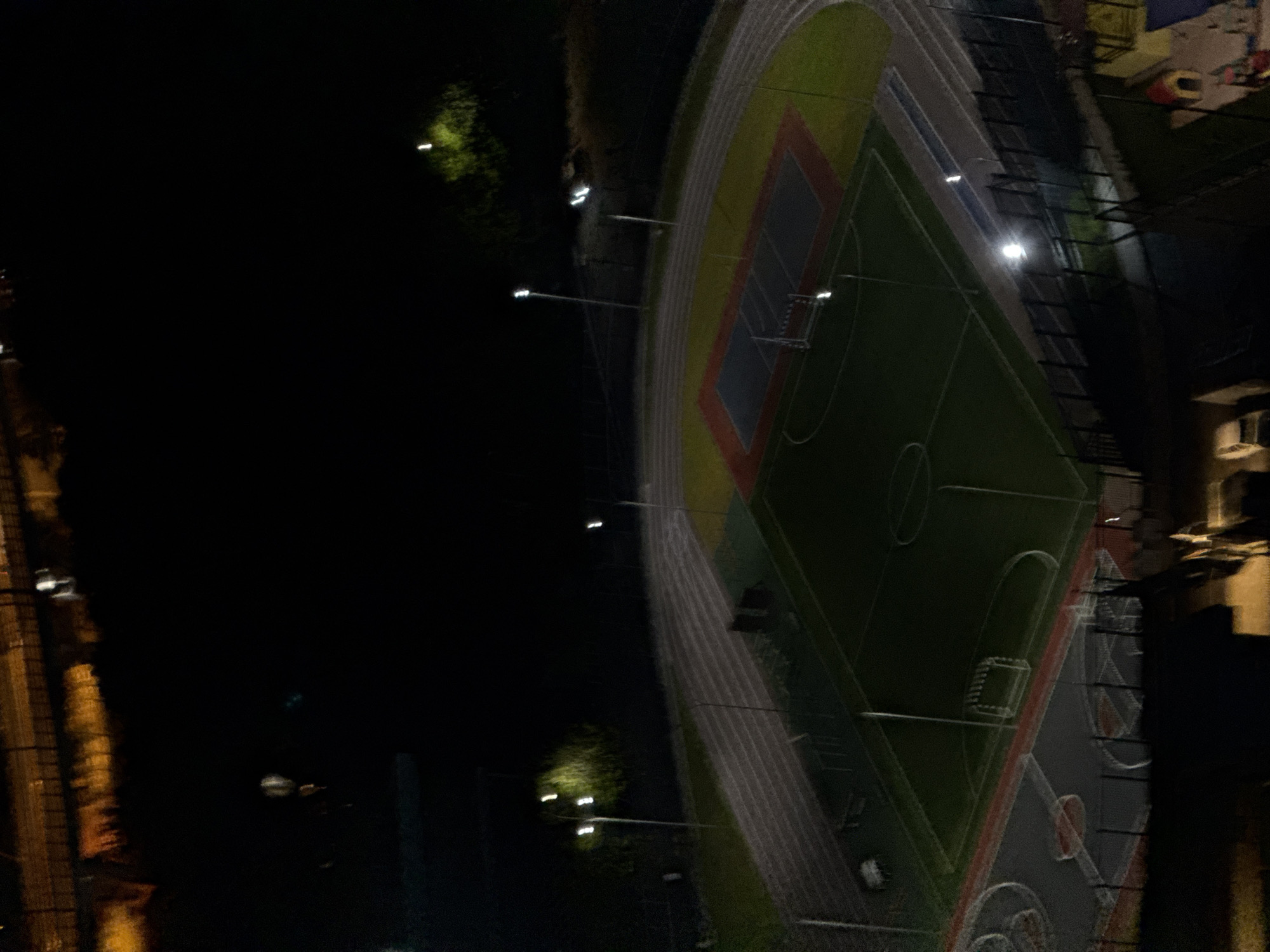 This is an aerial, night-time photograph of a sports complex in Sochi, Russia. The complex features a running track and a sports field, likely for soccer or football, illuminated by floodlights. The field is green with white markings, including goal areas. A basketball or volleyball court is also visible, with red and blue surfacing. The scene appears to be empty of people. The surrounding environment is dark, with some distant lights suggesting urban or suburban development. The photograph is grainy and somewhat out of focus due to the low light conditions.