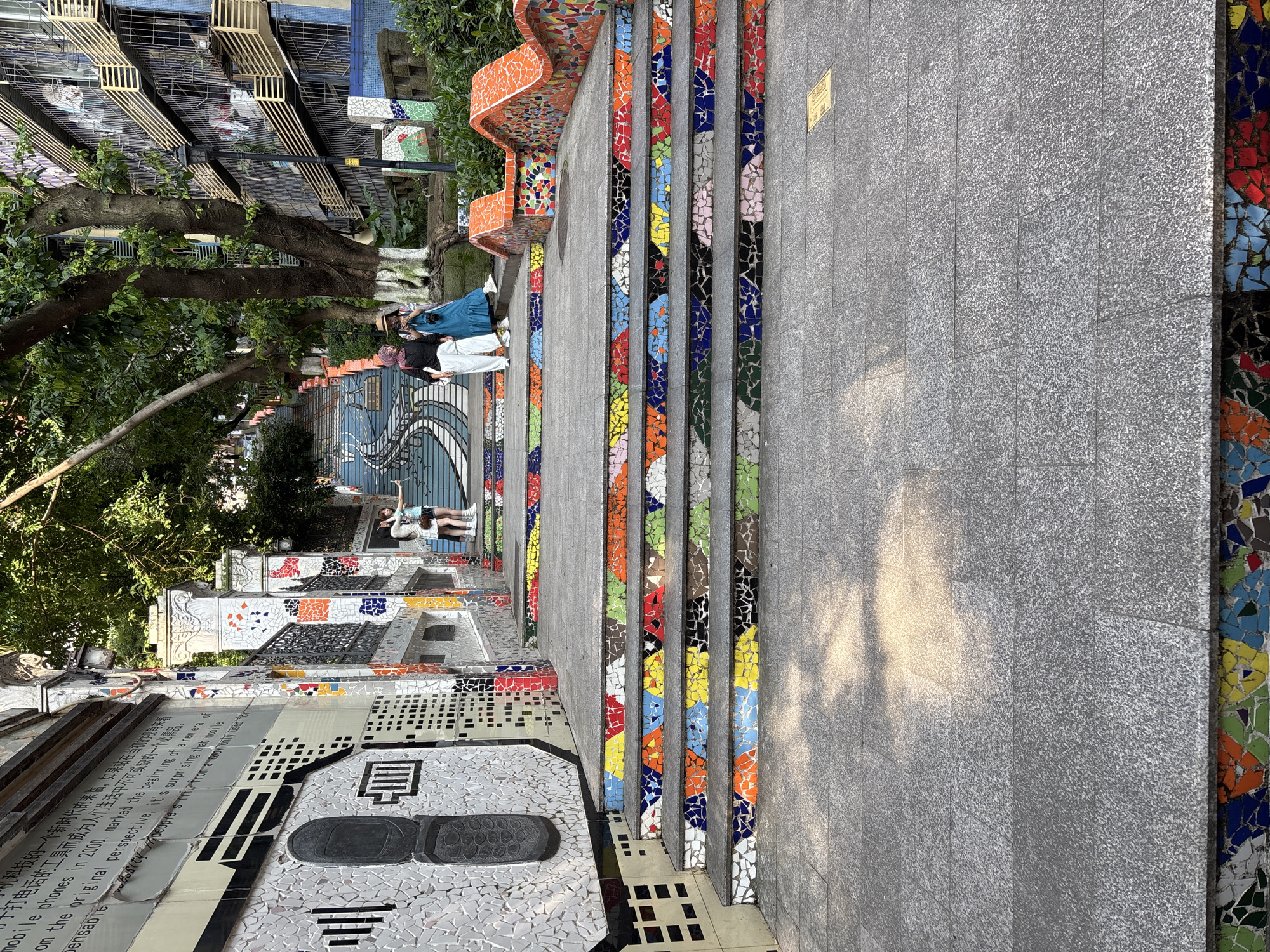 In Yuzhong District, China, a mosaic-tiled staircase is prominently featured, leading upwards. The staircase is adorned with vibrant, colorful mosaic tiles that form intricate patterns on each step's riser. The walls flanking the staircase are also decorated with mosaics, some featuring textual elements in Chinese and English, along with graphic designs.
In the mid-ground, three individuals are present. Two women are standing closer to the viewer, with one in a blue dress and a straw hat, and another in a hijab and white pants, both appearing to be looking at or taking photos. Further up the stairs, a third person is partially visible. Trees with lush green foliage are scattered along the sides of the staircase and above, suggesting an outdoor, urban environment. Behind the trees and buildings, apartment-like structures are visible. The ground in the foreground is paved with grey granite slabs, with sunlight dappling the surface. The overall scene captures a visually striking and artfully decorated public space.