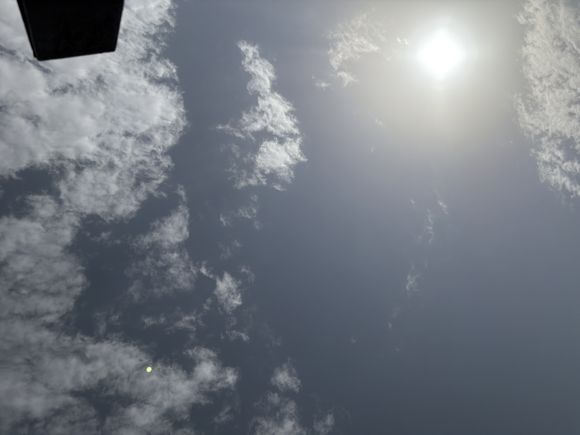 The image depicts a view of the sky, likely taken from Jos, Nigeria, on a partly cloudy day. The sun is a prominent, bright, square-shaped light source in the lower right quadrant, partially obscured by a haze of white clouds. Wispy white clouds are scattered across the predominantly greyish-blue sky. In the upper left quadrant, a small, bright, green circular object is visible, possibly a distant balloon or an artifact of the camera's lens. The upper right corner of the image is dominated by a dark, rectangular object, which might be part of a building or structure. The overall impression is of an outdoor scene with a focus on the atmospheric conditions.