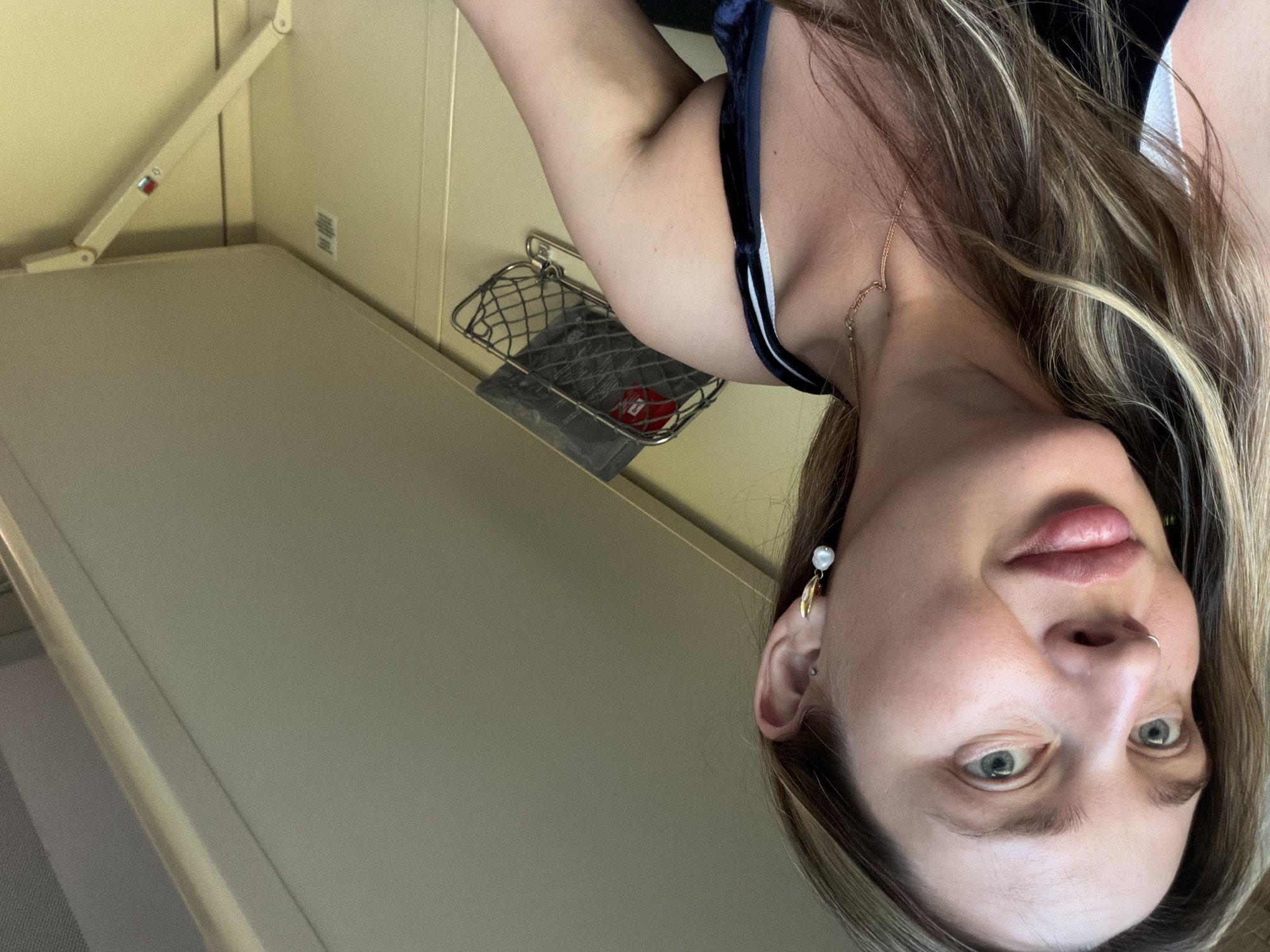 A woman with blonde hair is looking at the camera. She is wearing a blue top with a white stripe. She has a gold earring and a nose ring. The background is a light green wall with a metal rack. The location is not specified as Izhevsk, Russia, and therefore cannot be determined.