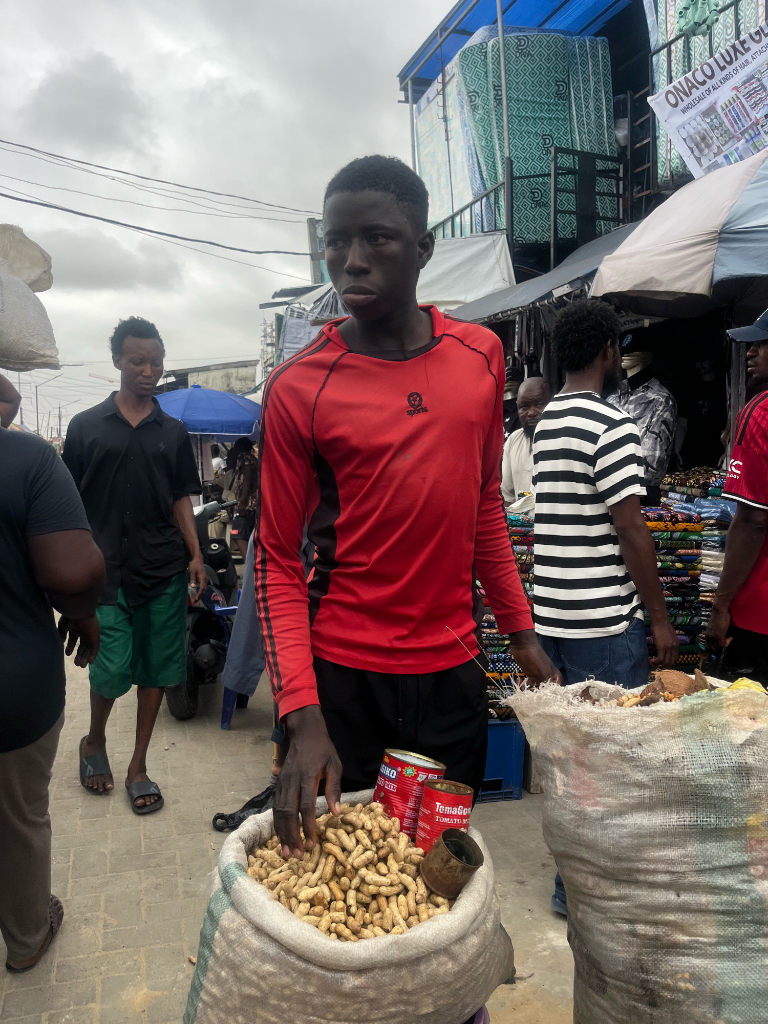 A street market scene in Lekki Phase I, Nigeria, features several people and numerous goods for sale. In the foreground, a young man wearing a red and black long-sleeved shirt stands near a large burlap sack filled with peanuts. Two red cans, one labeled "Temagol," are placed on top of the peanuts. To his left, another man in a black short-sleeved shirt and green shorts walks past. In the background, several stalls are visible, with one displaying stacks of colorful fabrics and another with mattresses. An overhead canopy structure with blue and white tarping covers part of the market. The ground is paved with interlocking bricks. The sky is overcast.
