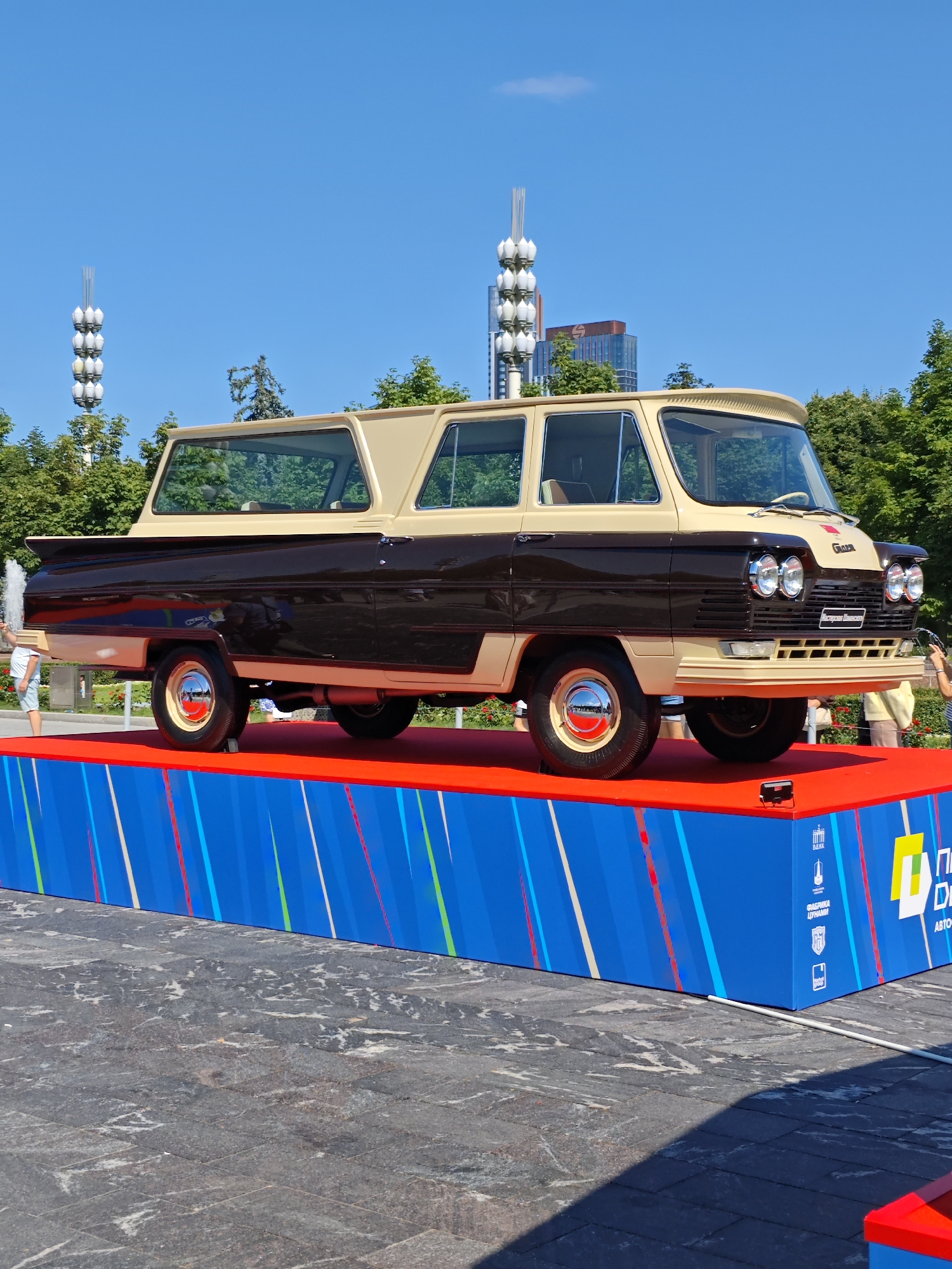 In Moscow, Russia, a vintage two-tone van, primarily brown with a cream-colored roof and accents, is displayed on a raised platform. The van, a Soviet-era GAZ-21 Volga station wagon variant, sits outdoors under a clear blue sky, surrounded by trees and modern buildings in the background. The platform is covered in red carpet on top, with blue sides adorned with colorful diagonal stripes and various logos and text, including Russian writing. A person in a white shirt and blue shorts is visible on the left side of the frame, near a fountain. Another person, wearing a yellow shirt, is seen on the right. The scene appears to be an outdoor exhibition or event, possibly related to automotive history.