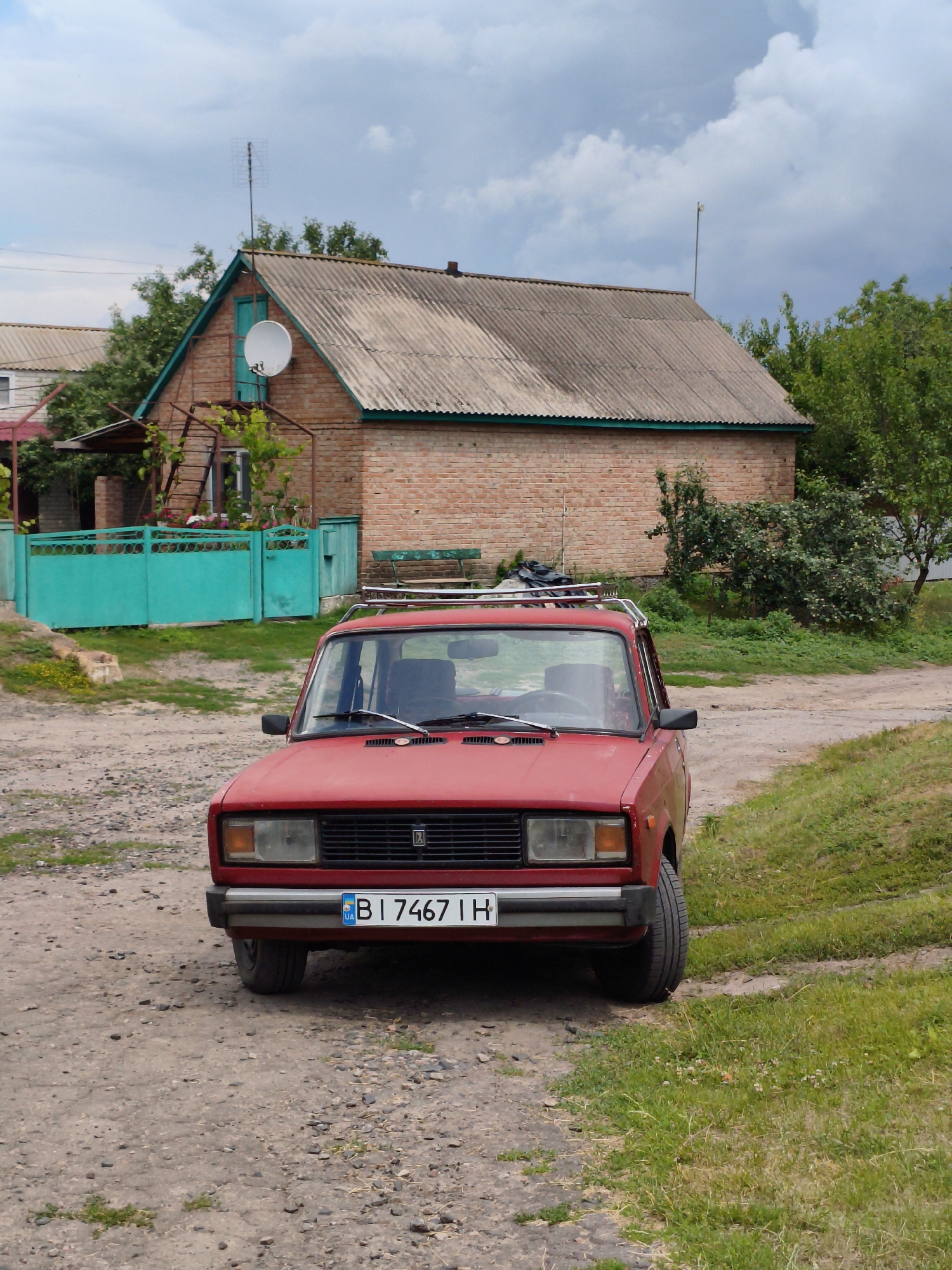 A red, older model sedan with a roof rack is parked on a dirt road in front of a brick house with a gray, corrugated roof. A turquoise fence is visible to the left of the house. The car's license plate reads "B I 7467 IH". Trees and green grass surround the scene, and the sky is partly cloudy. The location is identified as Ustyvytsia, Ukraine.