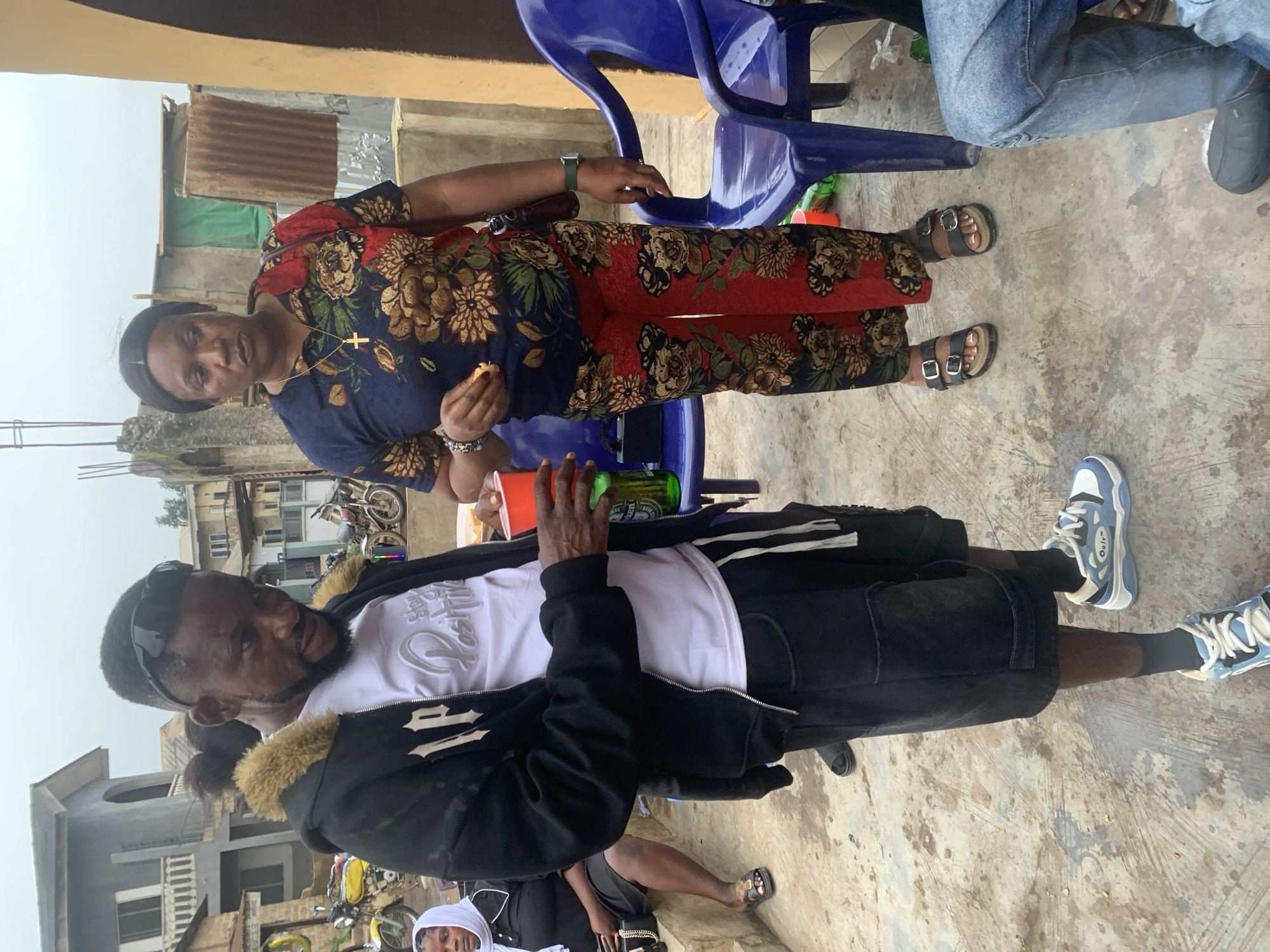 In Shagamu, Nigeria, on a cloudy day, a man in a black hooded jacket with a fur collar and a white t-shirt stands on a textured, dusty concrete surface. He is holding a red cup in his right hand and a green bottle in his left. He wears black shorts and light blue and white sneakers. A woman in a floral print blouse and red pants with a similar floral pattern stands to his right. She wears black sandals and a gold cross necklace. She holds a piece of food in her left hand. In the background, a woman in a white headscarf and black dress sits on concrete steps. Buildings, including a two-story structure and corrugated metal structures, are visible. Two motorcycles are parked on the left side of the image. A blue plastic chair is positioned behind the woman. Another person's legs, wearing blue jeans, are visible in the lower right corner. The overall scene suggests an outdoor gathering in a residential or commercial area.