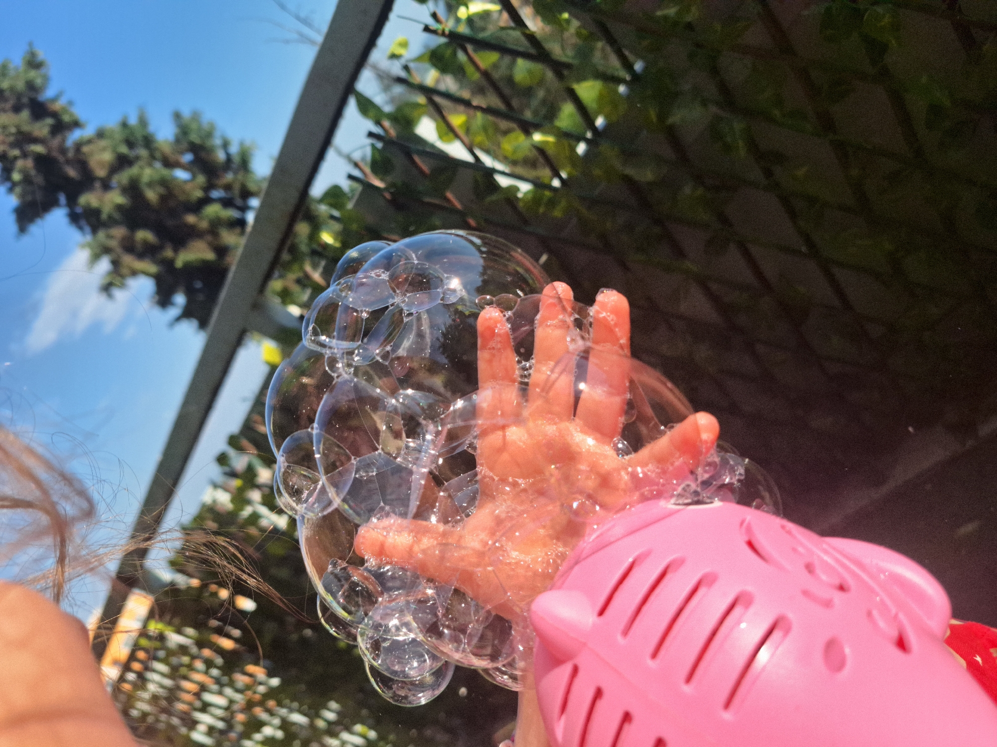 The image shows a child's hand reaching into a cluster of soap bubbles emanating from a pink, pig-shaped bubble machine. The hand is partially submerged in the bubbles, which are clustered tightly together. The bubble machine is in the lower right corner of the frame. The background includes a tree and the sky. The location is Unknown.