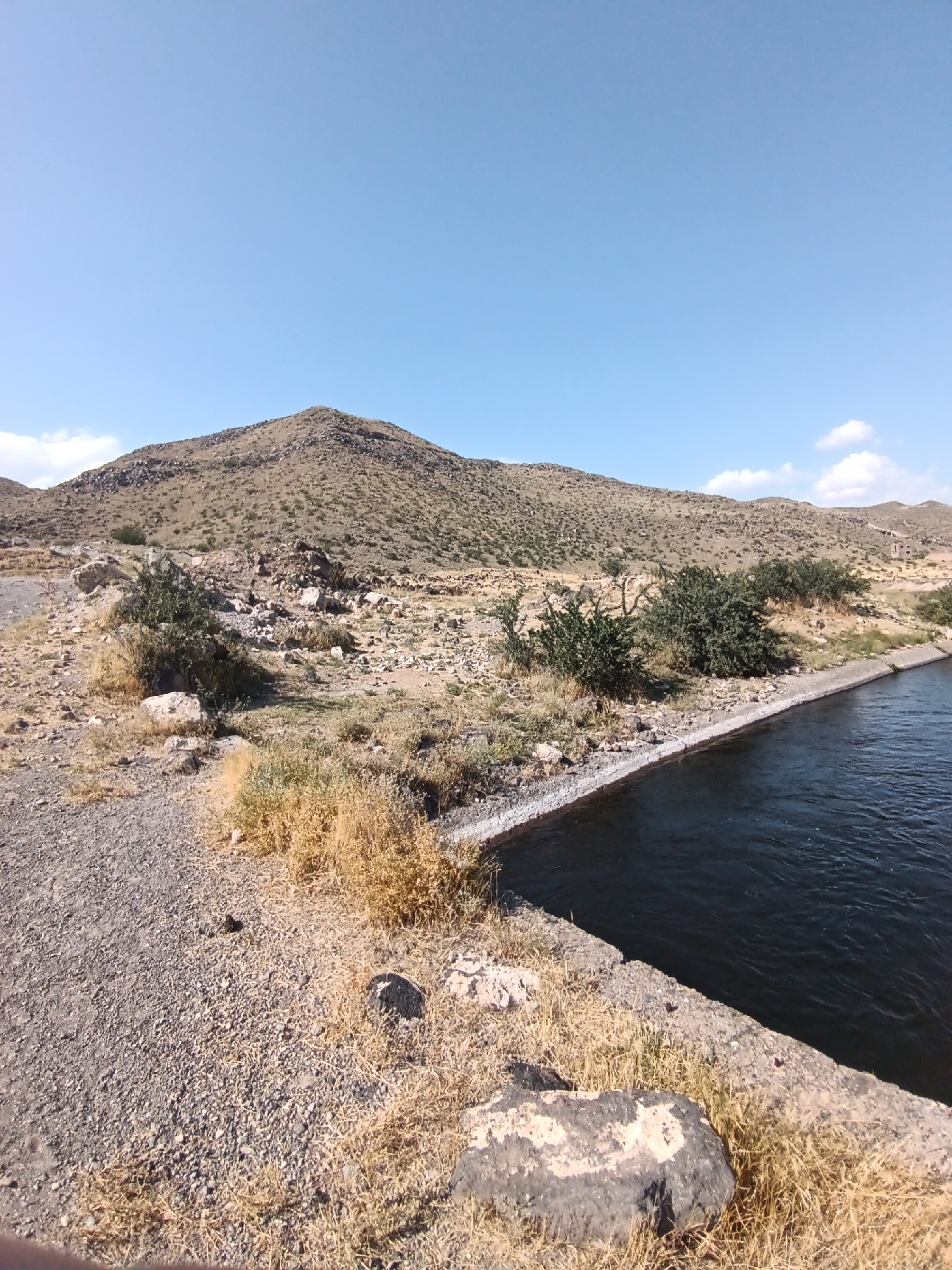 The image captures a section of a water canal running alongside a rugged, arid hillside under a clear blue sky. The canal, constructed with concrete walls, is filled with dark, rippling water. The hillside is covered with sparse, dry vegetation, rocks, and a few scattered green bushes. A gravel path borders the canal on the left side of the image. In the distance, a rounded, rocky hill dominates the horizon. The scene appears to be a rural or undeveloped area.