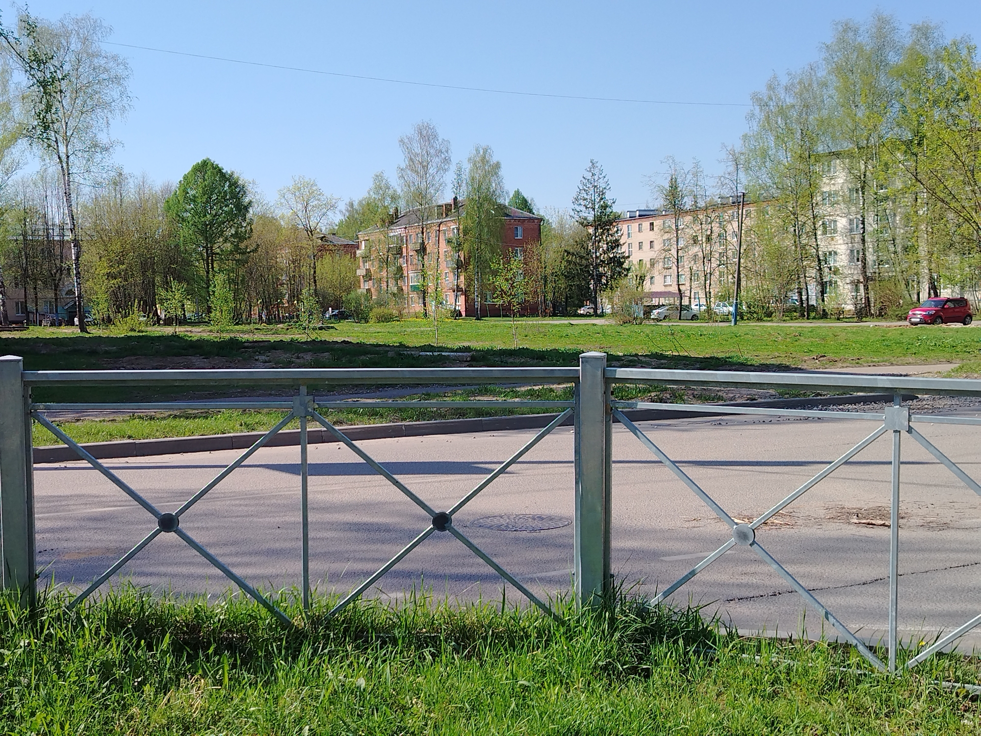 A street scene in an urban area of Unknown, Unknown is captured on a sunny day, with a focus on a grey metal fence that is partially obscured by green grass in the foreground. Beyond the fence, a grey asphalt road stretches across the scene, featuring a manhole cover. Across the road is a grass median, followed by buildings and trees in the background. The buildings appear to be apartments or low-rise structures, with brick and light-colored facades. Several vehicles are parked along the road adjacent to the buildings, including a red Kia Soul on the far right. The trees display fresh green foliage, indicating it may be springtime. The sky is clear and blue.