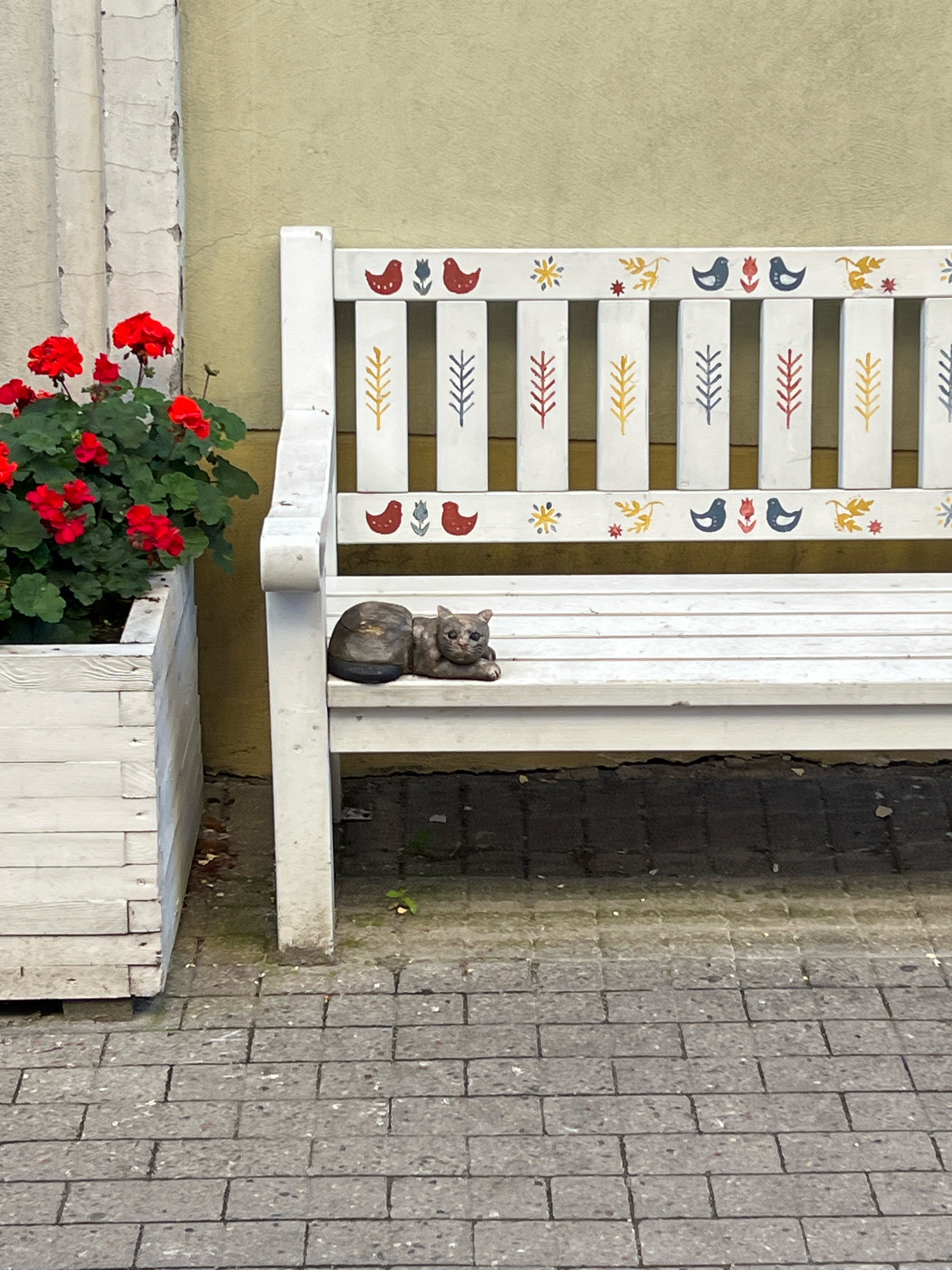 The image shows a white wooden bench decorated with colorful folk art patterns of birds and plants. A small, stone cat statue is lying on the bench. To the left of the bench is a white wooden planter filled with red geraniums. The scene is set on a cobblestone sidewalk in front of a building with a textured yellow wall. The overall setting suggests a charming outdoor space in a city. This image was captured in Vilnius, Lithuania.