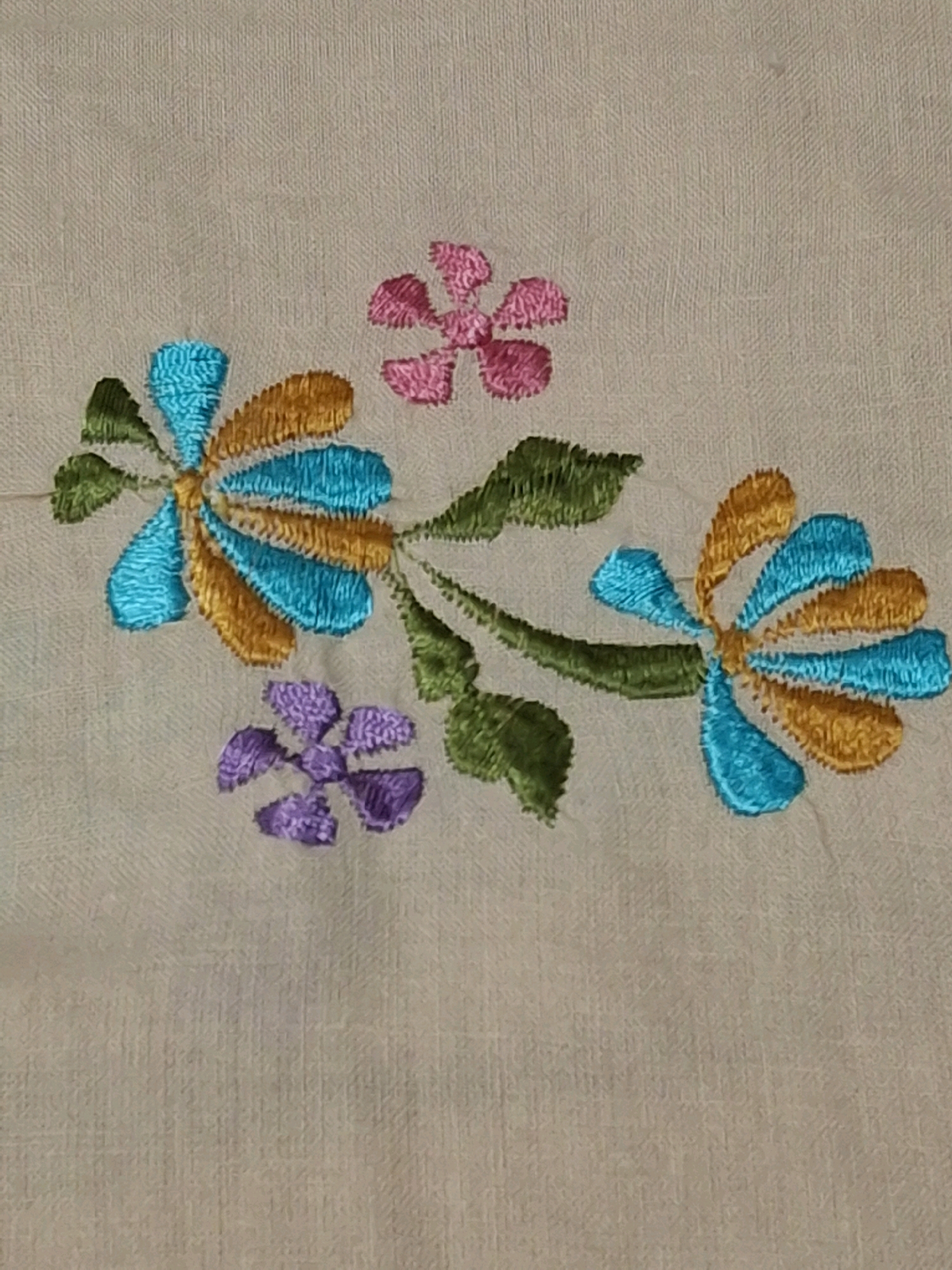 The image shows a close-up view of embroidered floral designs on a light beige fabric. The embroidery features several stylized flowers and leaves in vibrant colors including turquoise, gold, green, and purple. One larger embroidered flower is on the left, composed of alternating turquoise and gold petals radiating from a central point, with green leaves attached. Below this, a smaller purple flower with four distinct petals is embroidered. To the right, another larger flower with turquoise and gold petals is visible, connected by a stem with green leaves to the left cluster. A small pink flower with five petals is embroidered in the upper right portion of the image. The fabric appears to be a textured linen or similar material. The scene does not depict any specific location in Lumajang, Indonesia, but rather a detail of textile art.