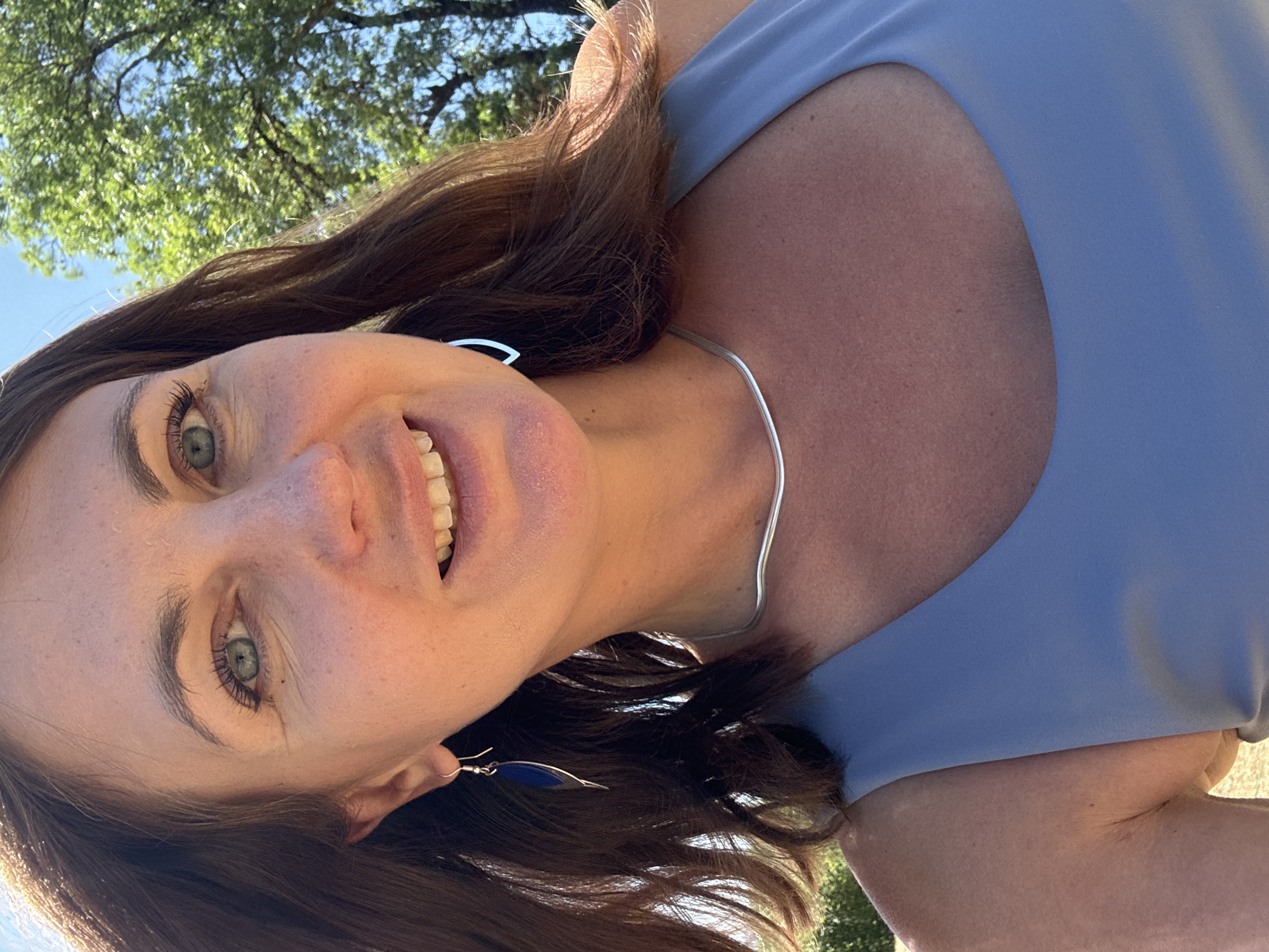 A woman with brown hair and blue eyes smiles at the camera in an outdoor setting. She is wearing a light blue tank top and a silver necklace. She also has on a pair of blue earrings. The background shows a tree with green leaves and a clear blue sky. The woman appears to be in the city of Coteaux du Lizon, France.