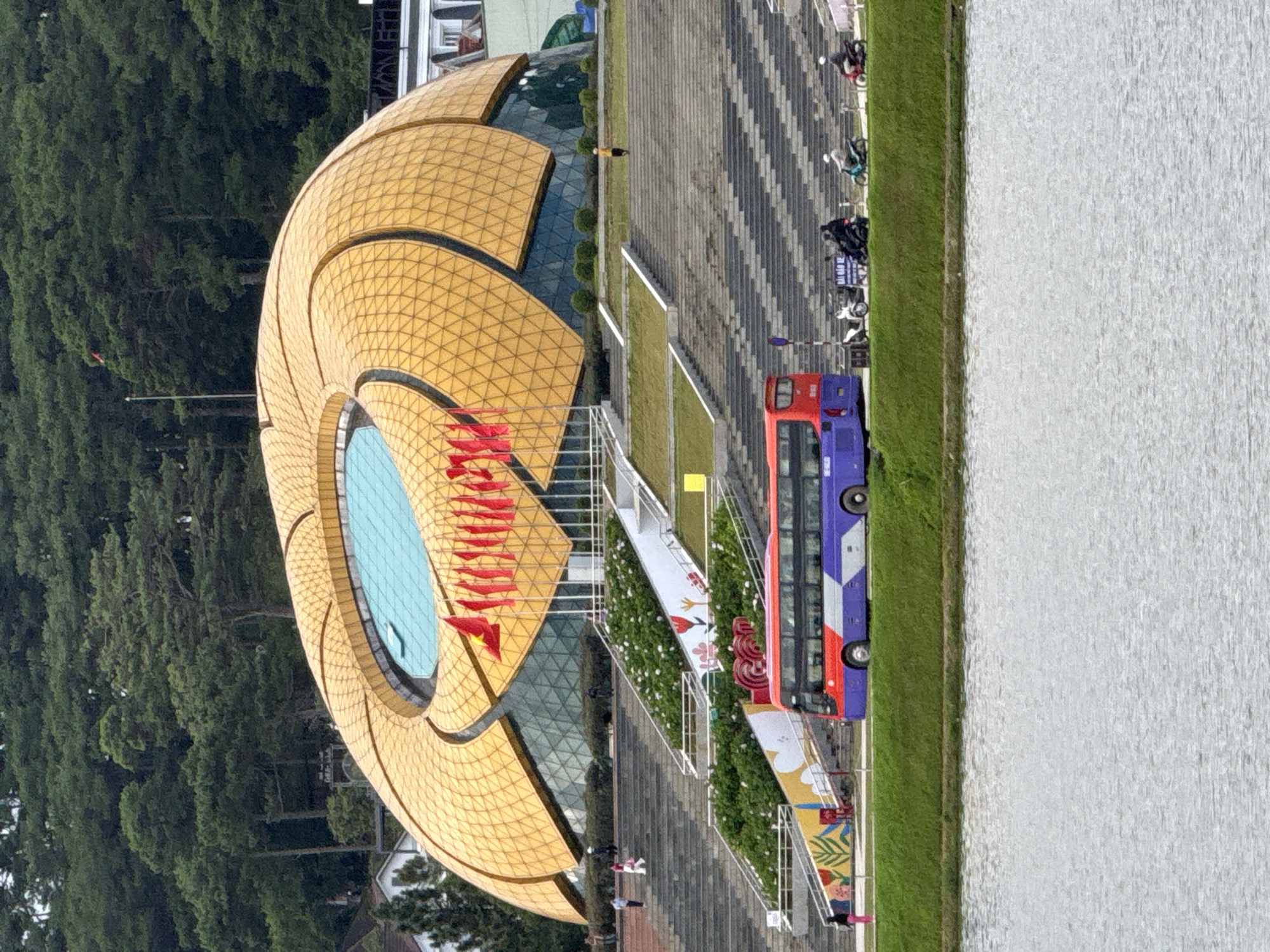 This image depicts the Da Lat Flower Garden, also known as the Da Lat Flower Park, located in Da Lat, Vietnam. The prominent structure in the background is the Da Lat Flower Garden Convention Center, characterized by its distinctive golden, petal-like roof and a large, circular blue skylight. Red flags are displayed along the front of the convention center.
In the middle ground, a wide, stone staircase leads up to the convention center, flanked by terraced gardens. The gardens feature green foliage and some white flowers. A large, double-decker bus, predominantly red and purple, is visible on the road in front of the gardens, with its doors closed. Several motorcycles are parked and being ridden on the road beside the bus.
The foreground is dominated by the calm surface of Xuan Huong Lake, with ripples indicating a slight disturbance on the water. The overall scene is set against a backdrop of lush green pine trees covering a hill behind the convention center. The lighting suggests it is daytime, possibly overcast.