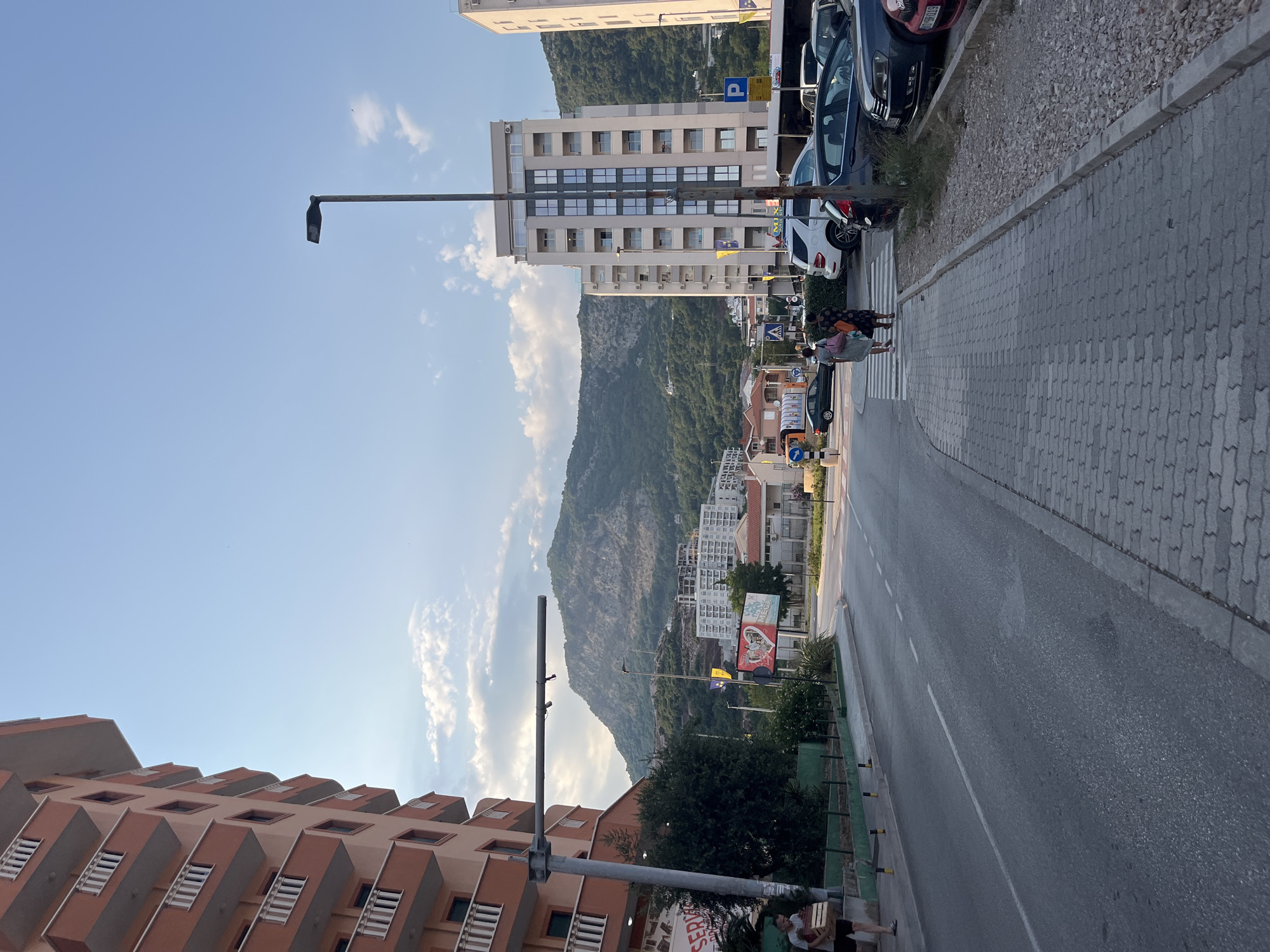 This is a vertical, eye-level shot of a street scene in Budva, Montenegro, at dusk. The sky is a clear blue with wispy white clouds. In the foreground, on the left, is a modern, multi-story building with a terracotta-colored facade and a repeating pattern of balconies. To the right, a paved sidewalk runs alongside a street. Several cars are parked along the sidewalk, and a few people are walking or standing. A pedestrian crossing is visible in the mid-ground.
Beyond the street, a large, green, tree-covered mountain dominates the background. Several buildings, varying in height and architectural style, are nestled at the base of the mountain and stretch up its slopes. A tall, modern building with a grid of windows stands out amongst them. Streetlights line the sidewalk.
Notable details include the time of day, suggested by the soft light and long shadows, indicating late afternoon or early evening. The weather appears to be pleasant, with a clear sky. The presence of palm trees and the architectural style of the buildings point to a Mediterranean or Balkan coastal location.
There is some visible text on a billboard-like structure in the mid-ground, but it is too blurry to read. A street sign with a "P" for parking is also visible. The overall mood of the image is peaceful and calm.