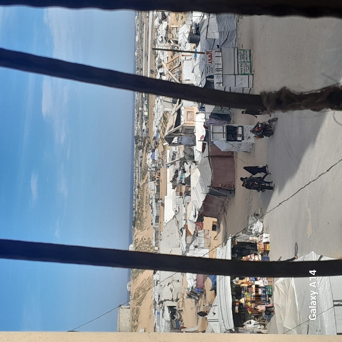 An elevated, slightly obstructed view overlooks a densely populated area with many temporary structures and dwellings. A road cuts through the scene, hosting pedestrians and a motorcycle. The structures are predominantly white or tan, likely shelters or simple homes. The backdrop reveals a bright sky and a distant horizon where the land meets the sea. The framing suggests the photo was taken from inside a building looking out.