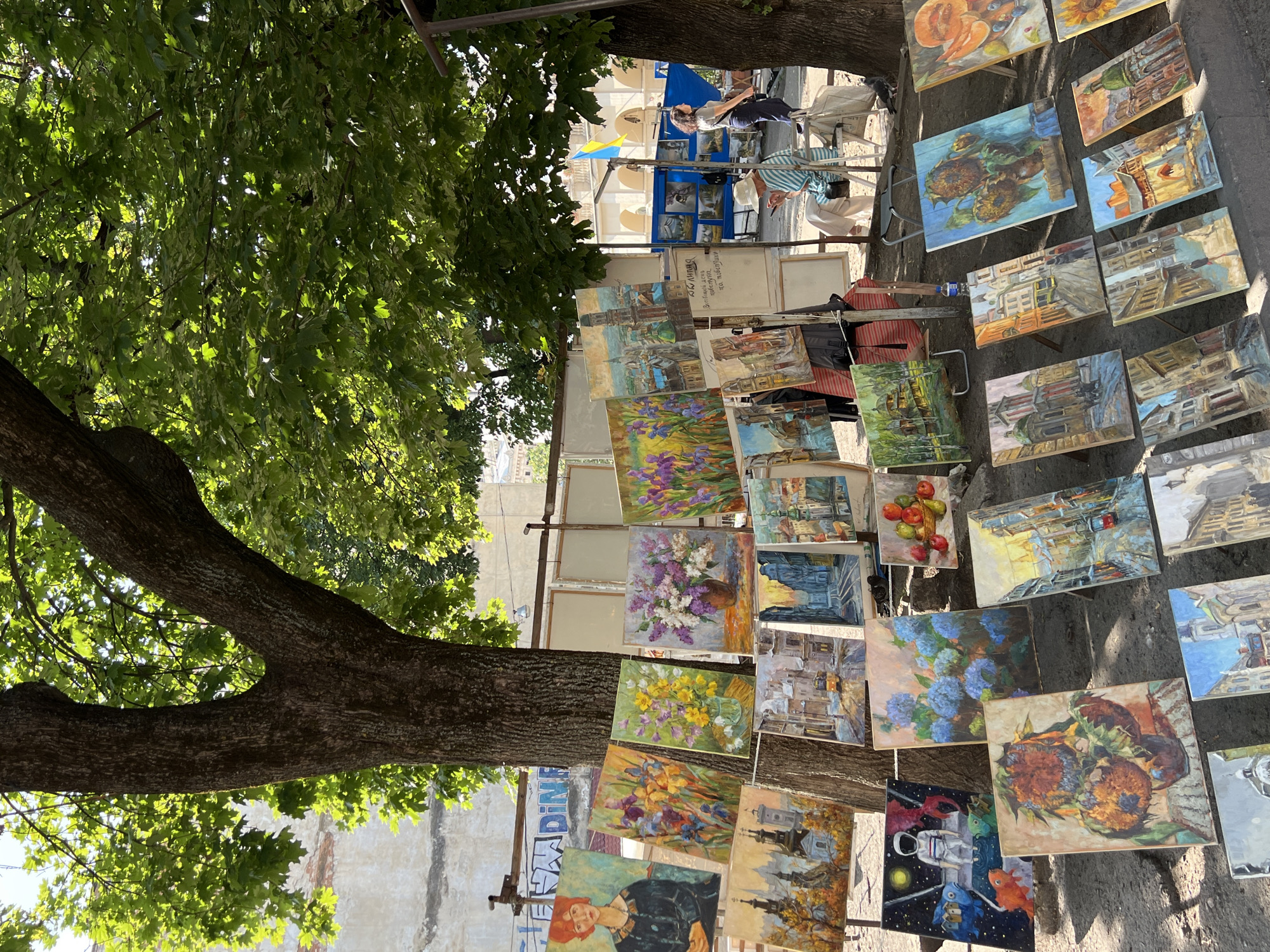 An outdoor art exhibition is displayed on a paved walkway in the city of Lviv, Ukraine, beneath the canopy of a large tree. Dozens of oil paintings, predominantly featuring cityscapes, floral arrangements, and portraits, are propped up on easels or hung from a makeshift display system. The paintings vary in size, with many depicting architectural scenes of Lviv's historic buildings, rendered in warm tones and with visible brushstrokes.
In the background, a building with arched windows is visible, and a Ukrainian flag flutters from a stand. Two people are present in the scene. A woman in a straw hat and striped shirt sits on a folding chair, looking down at her phone. Another woman stands near a display of paintings, looking towards the left of the frame. The ground is a mix of paving stones and dirt, with the base of the large tree dominating the center of the composition. Sunlight filters through the leaves of the tree, casting dappled shadows on the artworks and the ground. The overall impression is one of a vibrant, artistic marketplace set against the backdrop of a historic European city.