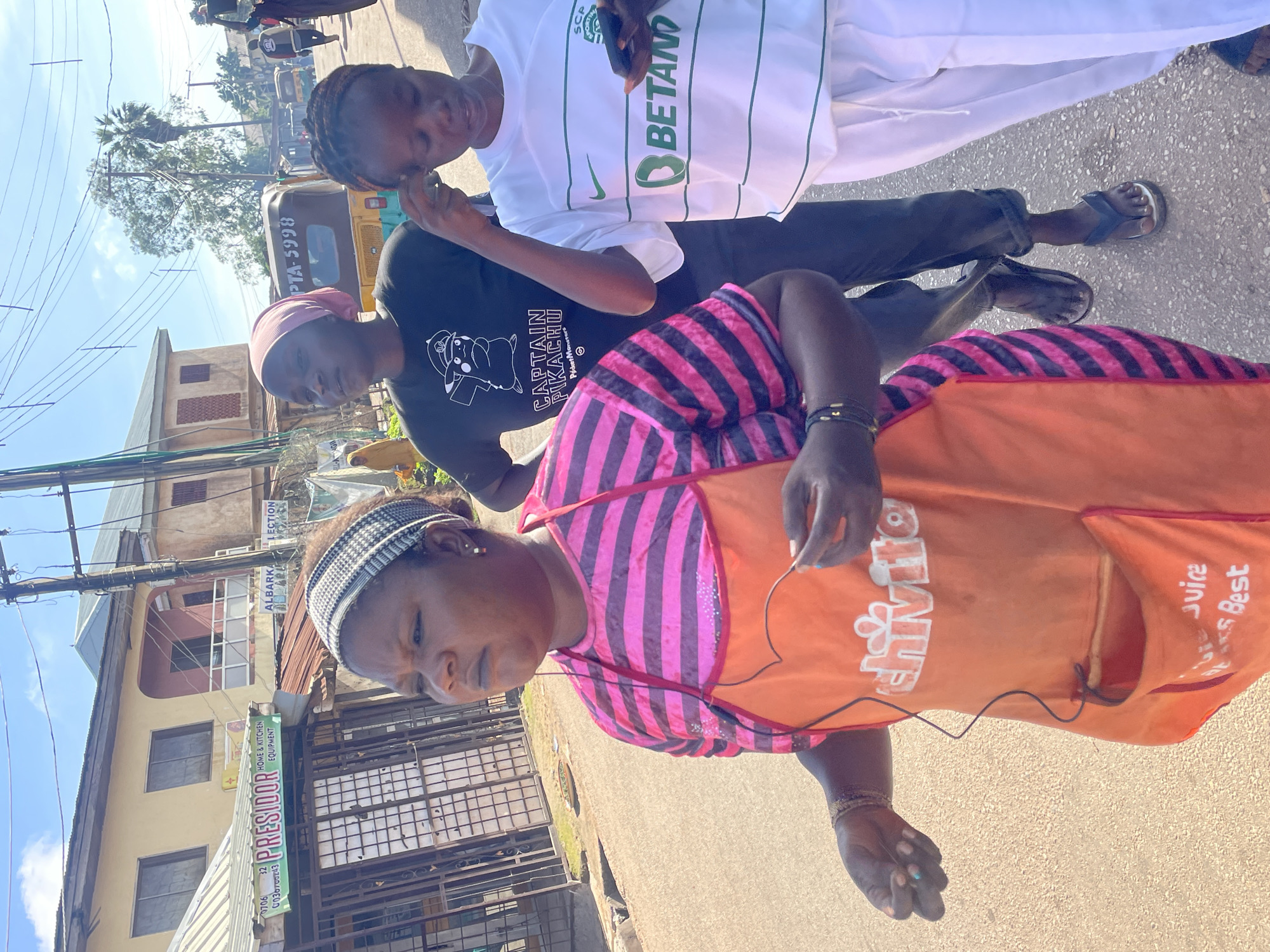 In Jos, Nigeria, on a paved street, three women are captured walking. The woman in the foreground, wearing a pink and purple striped shirt and an orange apron with the text "Chivite Juice Best," is looking to her left, holding what appears to be a wire or string. To her right, a woman in a black t-shirt with a "Captain Pikachu" graphic is looking forward. The third woman, on the far right, is wearing a white t-shirt with green accents and the "Betano" logo, and is looking down at her phone. Behind them, a yellow auto-rickshaw with the license plate "PTA-5998" is visible, along with other vehicles and buildings, suggesting a commercial area. The sky is bright and sunny, with utility poles and wires overhead.