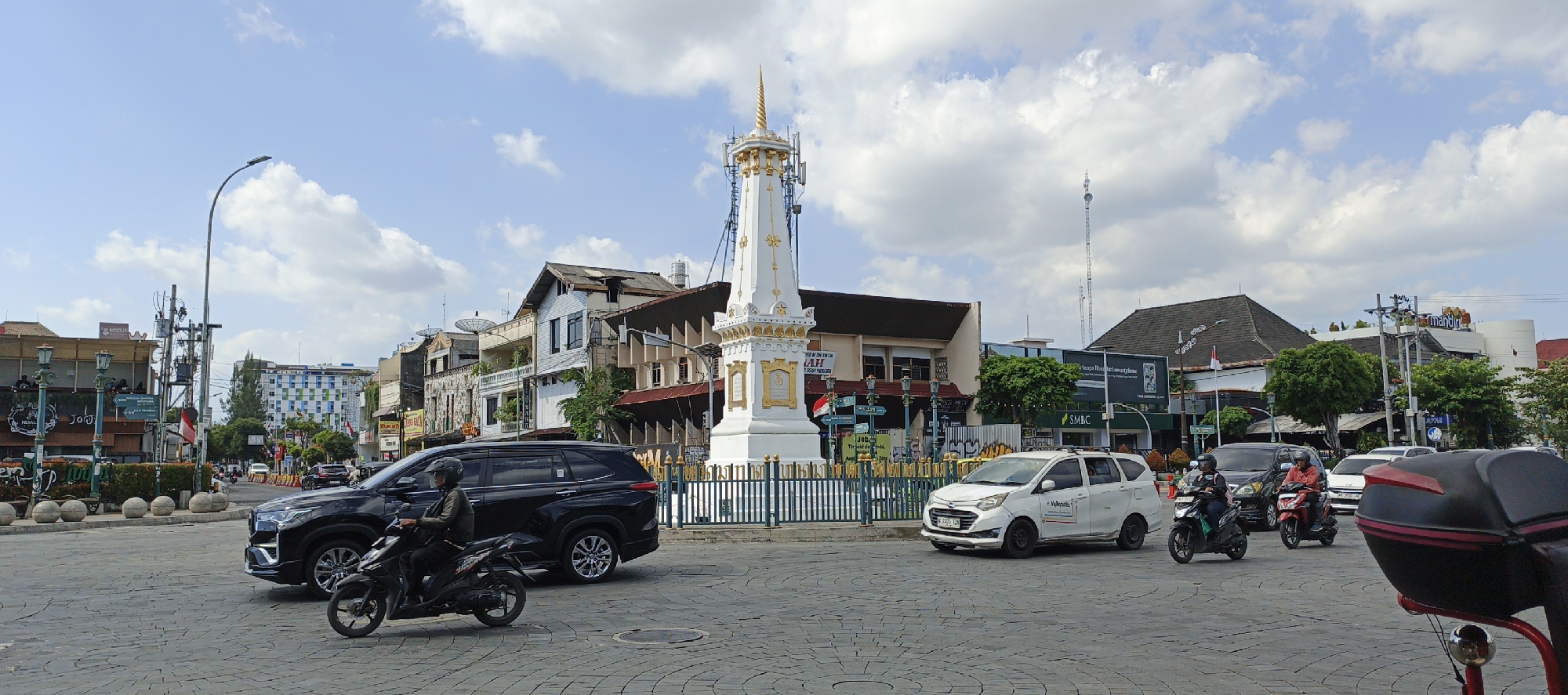 The image depicts a busy intersection in front of the Tugu Yogyakarta, a prominent monument in Yogyakarta, Indonesia. The monument, a white obelisk with golden ornate details and a spire, is centrally located in the frame. Surrounding the monument is a low, decorative metal fence.
On the left side of the image, a black SUV is stopped at the intersection, with a motorcycle rider in a black helmet and jacket in front of it, also stopped. The rider appears to be looking forward, possibly waiting for the traffic signal. In the middle ground, a white minivan is moving through the intersection, with a rider on a red motorcycle to its right. This rider is wearing a red shirt and a black helmet. Further back, another motorcycle rider on a dark motorcycle is also visible.
The background consists of a mix of buildings, including commercial establishments with signage, residential buildings, and a modern hotel with a distinctive grid-like facade. Trees are interspersed between the buildings. The sky is partly cloudy with white clouds against a blue background. The street is paved with interlocking bricks. The scene suggests a typical day with moderate traffic and pedestrian activity, captured from a street-level perspective.