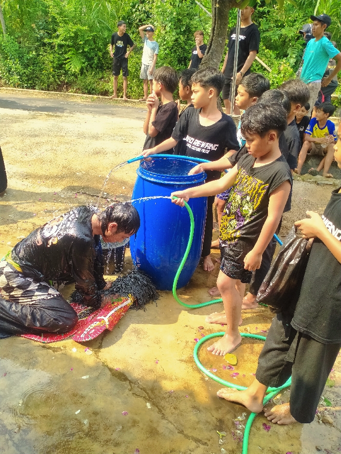 In Gebyog, Indonesia, a daytime outdoor scene depicts a cleansing ritual or preparation involving multiple individuals. A male subject, kneeling on wet, sandy ground, wears a black long-sleeved top with white patterning and dark patterned lower garments; his hair and clothing are thoroughly soaked as water is directed onto him from two hoses. Adjacent to him, a large, ornate object resembling a traditional mask or prop, featuring a red and white base with significant black, shaggy fiber elements, is also being rinsed with water.
Two young boys, barefoot, operate the hoses. One boy, wearing a black graphic t-shirt and black shorts, holds a green hose directing water towards the kneeling individual and the object. A blue hose, originating from a large, open-top blue plastic barrel filled with water, is held by another boy, partially obscured. The barrel serves as the primary water source.
Approximately ten other males, primarily juveniles and young adults, observe the activity. Some stand near the blue barrel, wearing black t-shirts, one visible with "JAGO REWANG CATURE REJO" text. Others are positioned further back along a dirt path bordering a paved road, with a backdrop of dense green foliage and trees. Their attire includes various t-shirts (black, light blue, teal, yellow-blue) and shorts.
The immediate ground is wet and muddy, with scattered purple and pink flower petals. A small yellow object lies on the ground near the green hose. Bright, unfiltered daylight illuminates the entire scene.