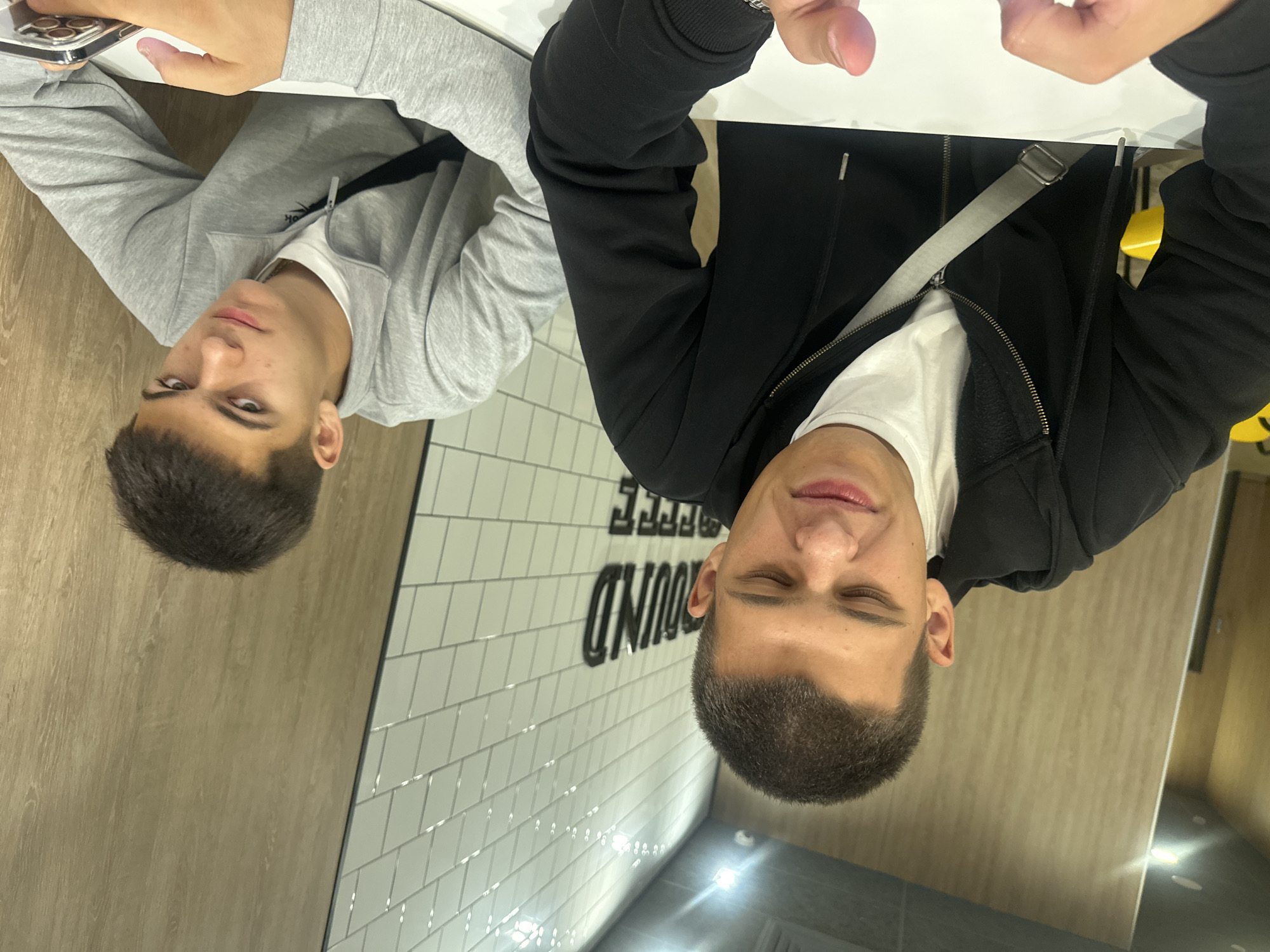 Two young males are seated at a white table in what appears to be a cafe or restaurant. The person on the left, closer to the camera, is wearing a black zip-up hoodie over a white t-shirt and has a grey strap visible across their chest. They are looking down with their eyes partially closed and their mouth slightly open. The person on the right is wearing a grey zip-up sweatshirt over a white t-shirt and is holding a smartphone in their right hand. They are looking directly at the camera with a neutral expression. Behind them, a wall is decorated with white subway tiles and a wooden panel. The overall lighting suggests an indoor setting, possibly a commercial establishment. There is no visible indication that this scene is located in Birobidzhan, Russia.
