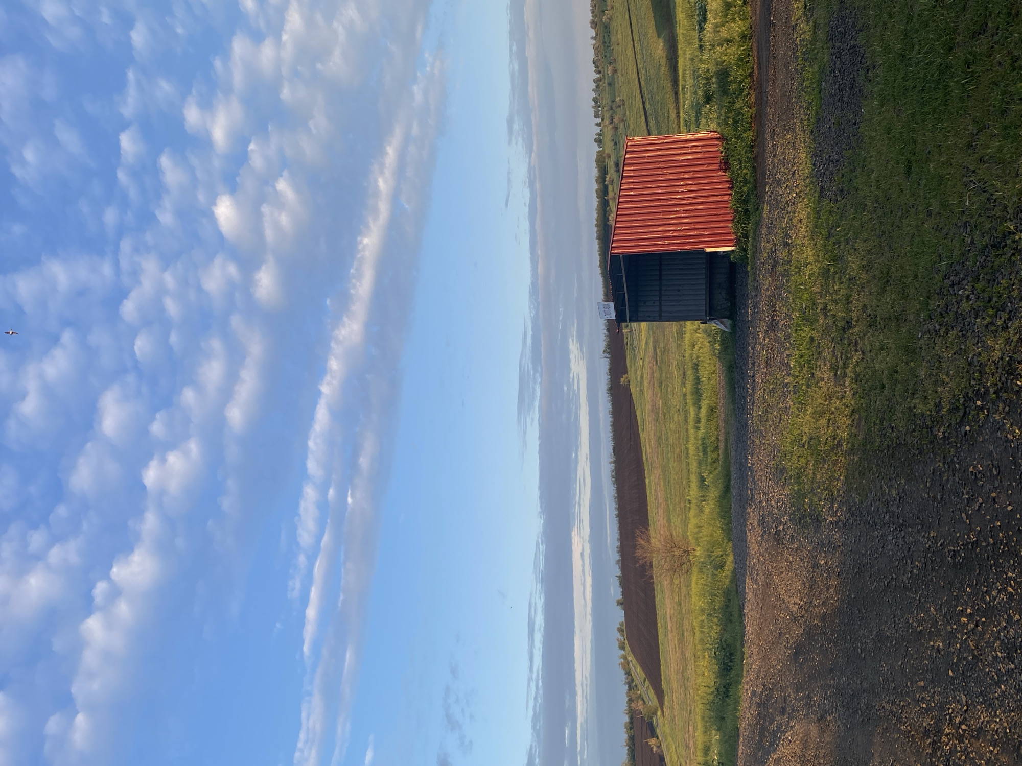 The image depicts a landscape with a building, field, and sky.
The building is small and rectangular, with a red roof and dark walls. It sits on the edge of a field and a gravel road.
The field is green with growing crops.
The sky is blue with scattered clouds.
A small bird is seen on the upper left of the sky.
Location: Unknown, Unknown.