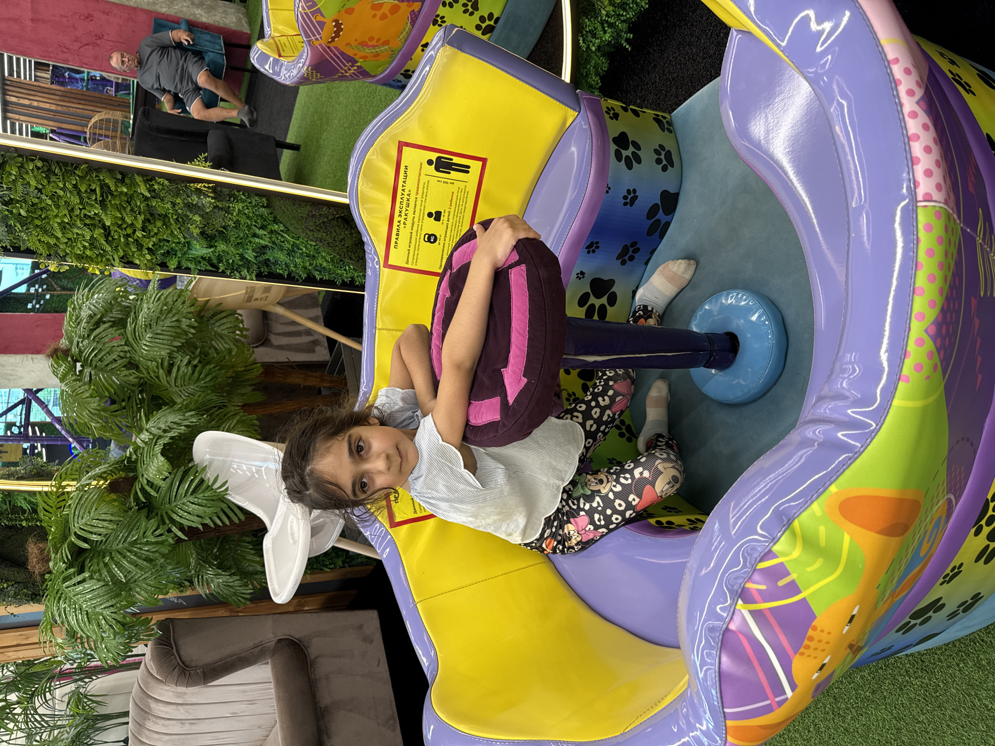 A young girl with dark hair and brown eyes is lying down on a colorful, plush toy. She is wearing a light-colored, striped t-shirt and patterned leggings. Her arms are wrapped around a large, dark purple cushion with pink stripes. The toy she is on is a large, shell-shaped seat, predominantly yellow and purple, with green and pink accents. The seat is decorated with paw prints and cartoonish animal graphics.
Behind the girl, a man in a grey t-shirt and shorts is sitting on a dark blue chair. He is looking towards the camera. The background suggests an indoor play area or entertainment venue. There are artificial plants with large green leaves visible, and a plush grey sofa is also in the background. The floor appears to be covered with green artificial turf.
A sign with text in Russian is visible on the yellow part of the toy. The text includes "ПРАВИЛА ЭКСПЛУАТАЦИИ" (Rules of Operation) and indicates that the module is a "RAKUSCHKA" (Seashell) and is a trauma hazard. It also provides age and height restrictions, stating it's for children up to 150 cm tall and can accommodate up to 2 children.
The lighting suggests an indoor, well-lit environment. The overall mood seems relaxed and playful, with the girl appearing comfortable and engaged with the toy. The scene is set in what appears to be a family-friendly entertainment space.