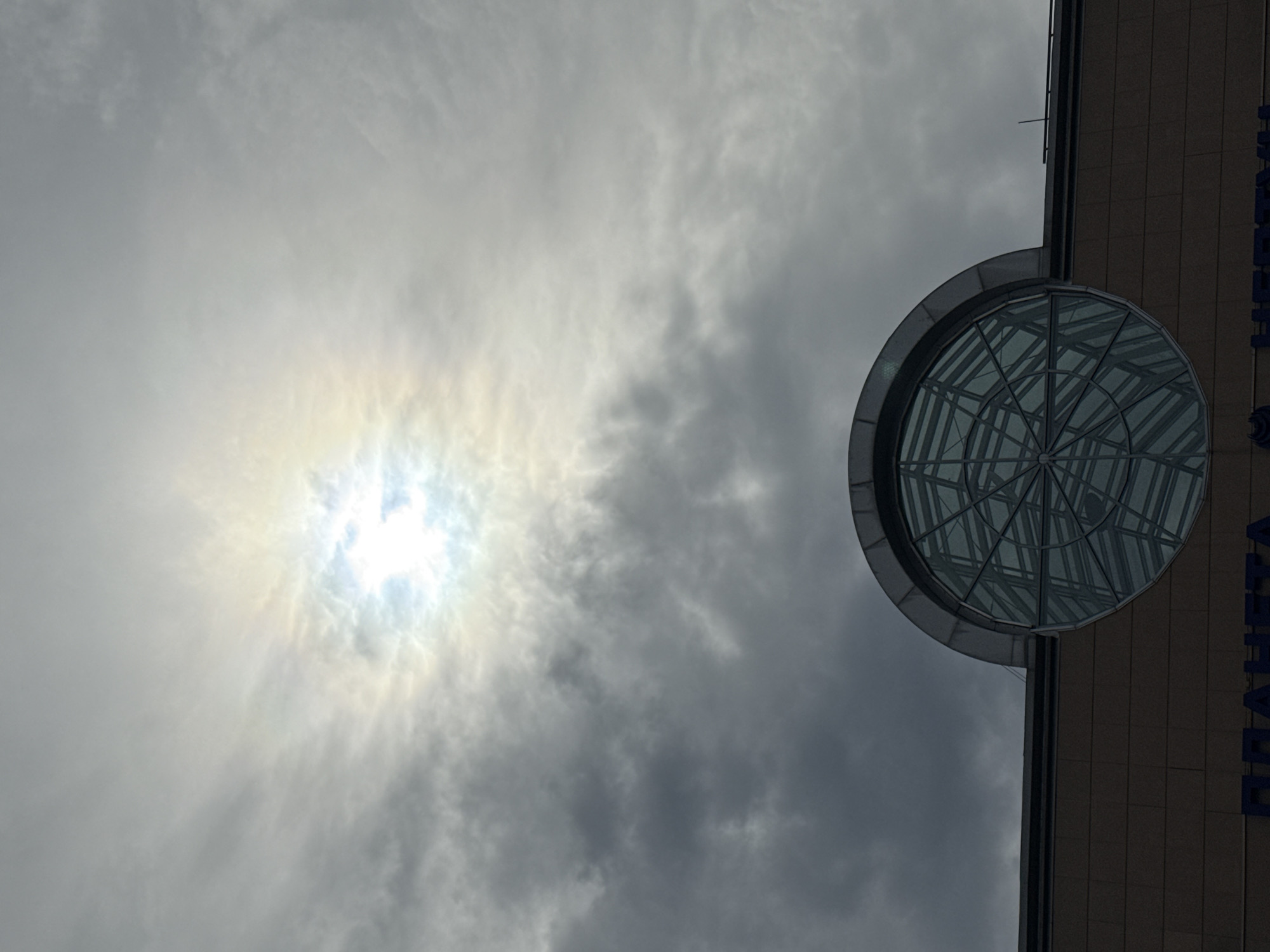 The image shows a cloudy sky with the sun partially obscured by clouds, casting a bright light. To the right, a portion of a building is visible, featuring a circular glass dome atop a facade. The building's name, "ПЛАНЕТА НЕПТУН," is partially visible in Cyrillic script, indicating its location in Russia. Based on the building name, this is likely the "Planet Neptune" oceanarium located in Saint Petersburg, Russia.