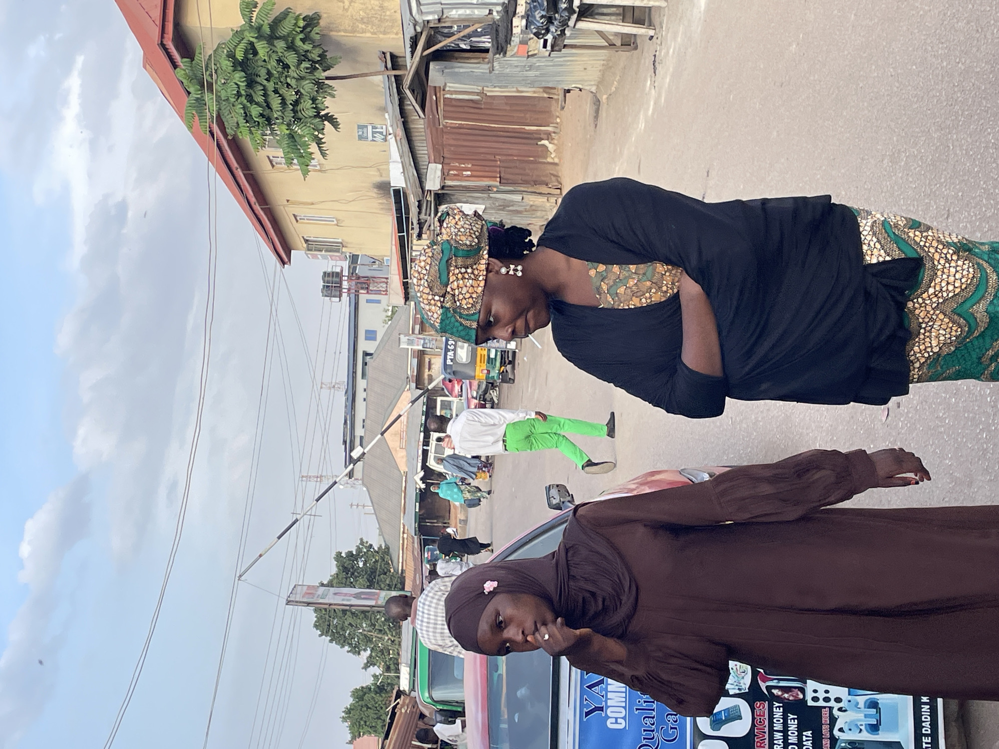 The image captures a street scene in Jos, Nigeria. Two women are prominently featured in the foreground. One is wearing a brown hijab and dress, while the other is in a black top and a patterned head wrap. The street is bustling with activity, showing other people walking and vehicles passing by. In the background, buildings and shops line the street, reflecting a typical urban environment in Jos. The signage on a car suggests services related to money transfer and data.