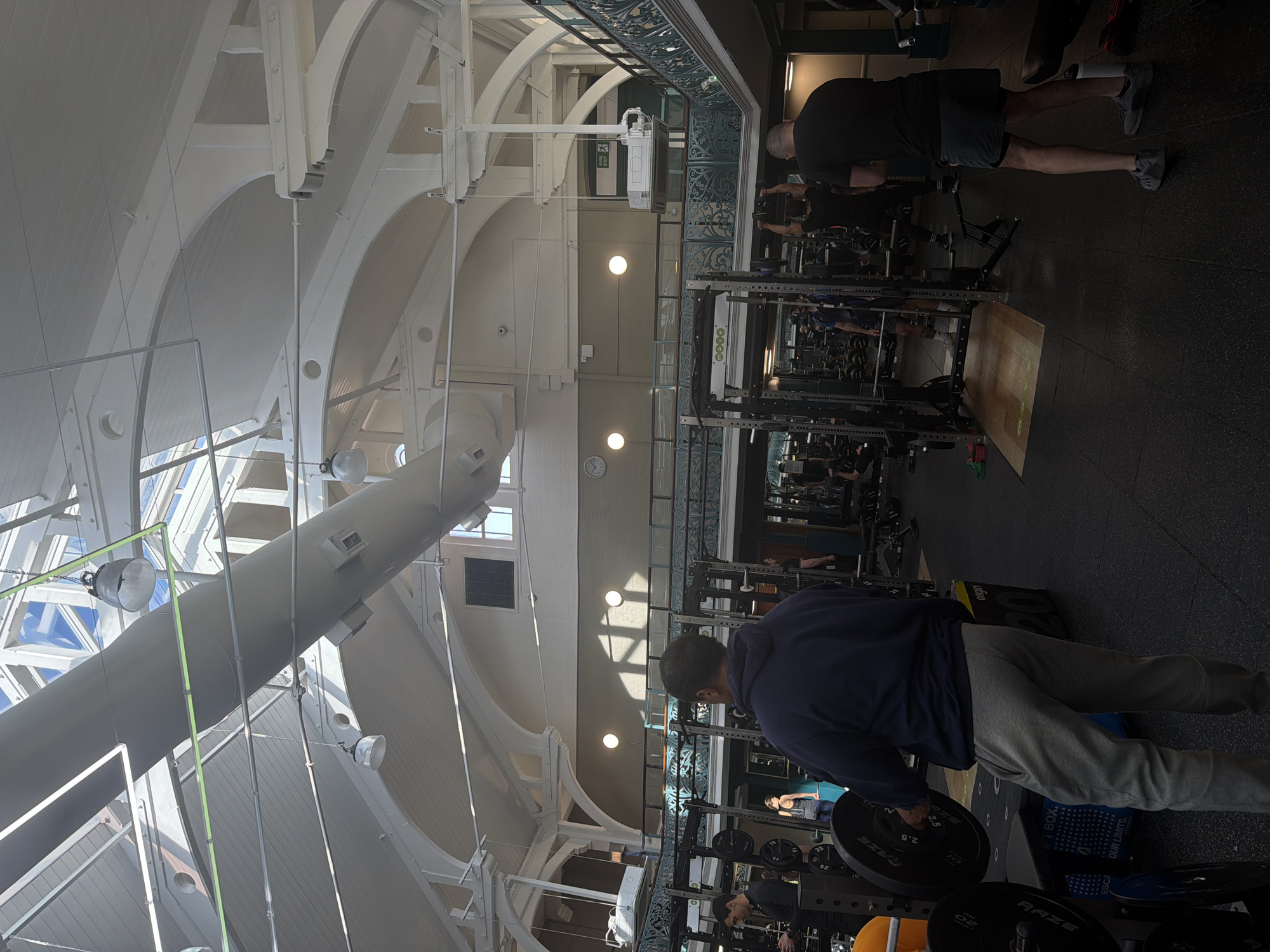 The image captures the interior of a fitness gym in the city of London, United Kingdom. The gym's architectural design features a high, arched ceiling with exposed beams and supports, giving it an airy and spacious feel. Natural light filters through windows in the roof, supplementing the artificial lighting provided by round overhead lights.
Several individuals are engaged in various exercises. In the foreground, a person is lifting weights, wearing a navy blue sweater and gray sweatpants. In the background, other people are working out using the gym's equipment, including weight racks and benches. A man in a black shirt and shorts is standing near a weight rack.
The gym is equipped with a variety of weightlifting and fitness machines. A second level balcony overlooks the gym floor. The floor is covered in dark-colored gym flooring, and there are weight plates and other fitness accessories scattered throughout the space.