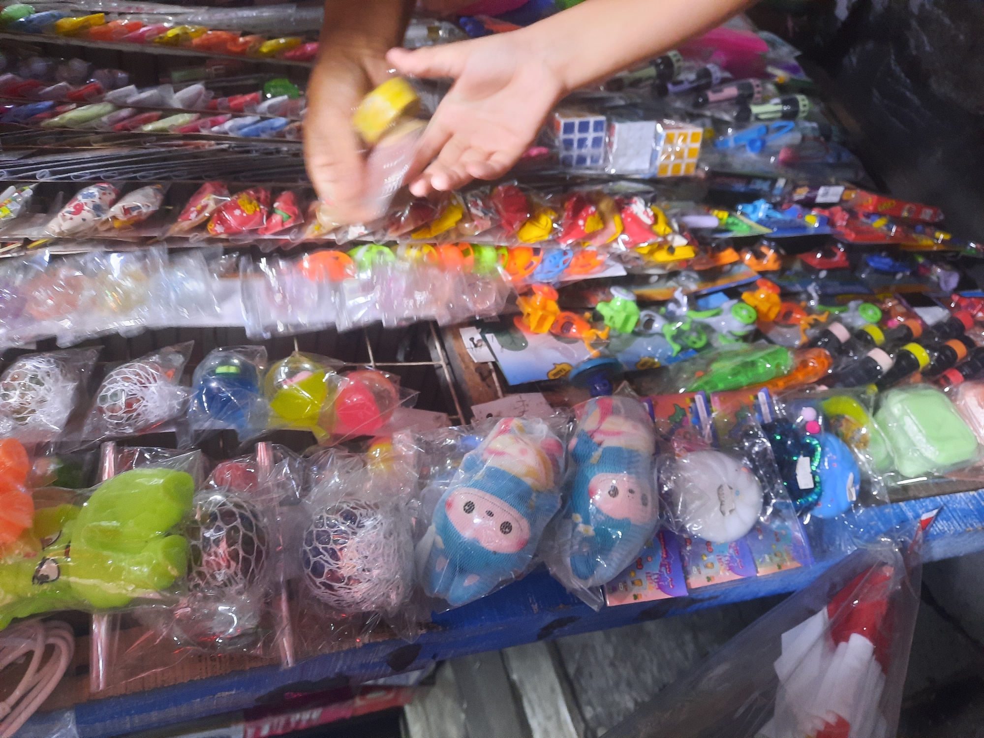 The image captures a close-up view of a stall displaying a variety of small toys and trinkets, likely a street vendor setup. The stall appears to be located outdoors in North Jakarta, Indonesia, suggested by the dense arrangement of items, the visible textures of plastic packaging, and the hint of a weathered wooden structure supporting the display.
Dominating the left side of the frame are rows of toys packaged in clear plastic. Several of these are stress balls, some with nets revealing internal textures, and others are soft, squishy figures with cartoonish faces, resembling dolls or plush toys. In the foreground, a child's hands are visible, one holding a yellow object, possibly a small toy or candy, and the other hand reaching towards it. This interaction suggests a customer or a child browsing or purchasing items.
The stall is packed with an assortment of colorful toys. Hanging vertically are numerous small, individually packaged items, many with bright, varied colors like red, yellow, green, blue, and orange. These include what appear to be miniature puzzle cubes, small plastic figures, and other novelty items. The overall impression is one of a busy marketplace or a roadside stall overflowing with merchandise. The background is slightly blurred, but it implies a narrow street or walkway, typical of many bustling commercial areas in North Jakarta. The lighting suggests it might be daytime, with some artificial light possibly contributing to the scene's brightness.