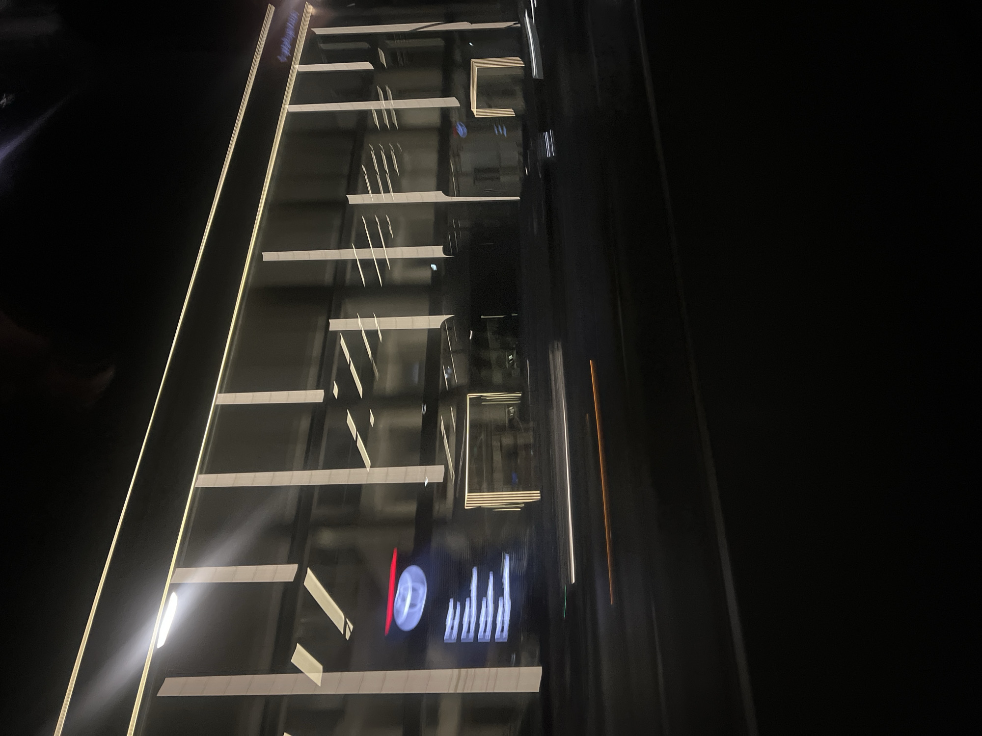 The image shows a blurred, nighttime view of a modern building facade with horizontal and vertical lighting elements. The facade appears to be made of glass and has a sleek, contemporary design. The lighting is predominantly white and yellow, creating a dramatic effect against the dark background. Some of the lighting elements are vertical, resembling illuminated pillars, while others are horizontal stripes. There is a prominent red circular logo with white text on the left side of the building. The image is taken from a moving vehicle, as indicated by the motion blur in the foreground, which shows streaks of light from streetlights or headlights. The overall impression is one of urban dynamism and modern architecture.