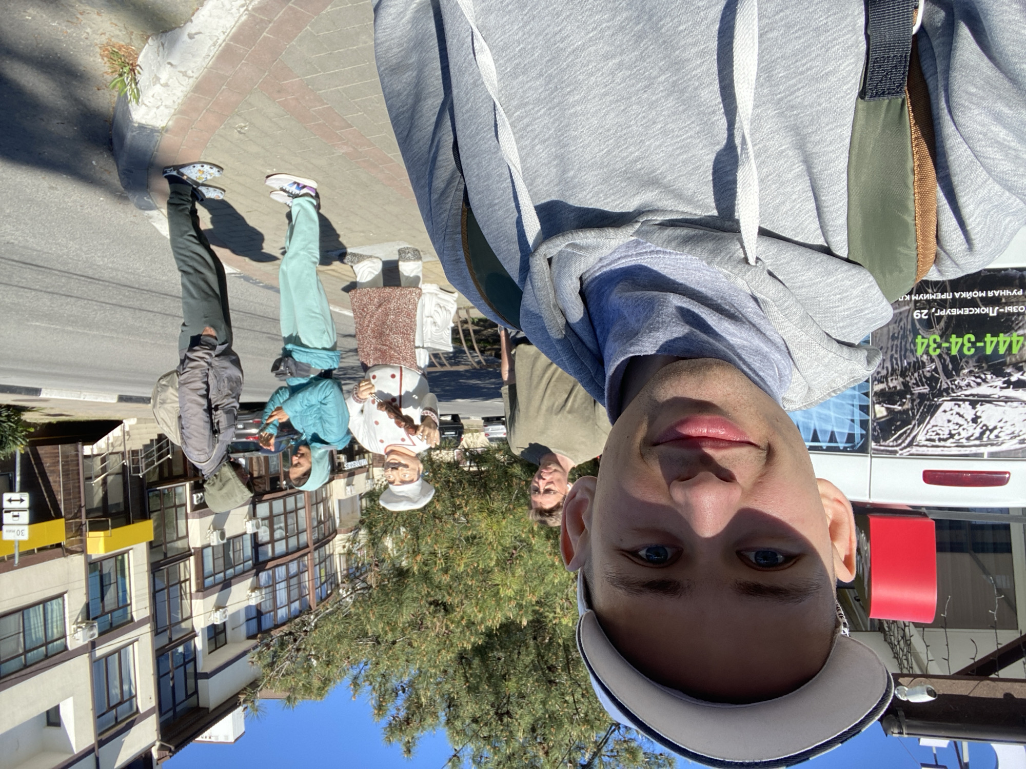 A selfie captures a bright, sunny street scene in Gelendzhik, Russia. A young man, wearing a grey hoodie and a white and blue baseball cap, smiles subtly into the camera, his blue eyes prominent. He occupies the foreground, with the street and surrounding environment stretching out behind him.
In the mid-ground, two individuals walk on a paved sidewalk. One wears a dark jacket and pants, and the other a light blue jacket and lighter pants. Further back and to their right stand two human-sized statues of chefs; one is clearly visible in a white uniform with a red and white apron, holding a skewer. A man with glasses wearing an olive-green top is also visible behind the statues.
The background features multi-story buildings with numerous windows lining the left side of the street. Lush green trees and bushes, including a prominent pine-like tree, add natural elements to the urban setting. On the right, a white van or bus is partially visible, bearing Cyrillic text that includes a phone number "444-34-34" and what appears to be an advertisement for a "РУЧНАЯ МОЙКА ПРЕМИУМ КЛАССА" (Premium Manual Car Wash) at "РОЗЫ-ЛЮКСЕМБУРГ, 29" (Rosa Luxemburg, 29). A red container is next to the van. A street sign with "30 мин" (30 min) and two opposing arrows suggests a time-limited zone. The clear sky and strong shadows indicate it is daytime with good weather.