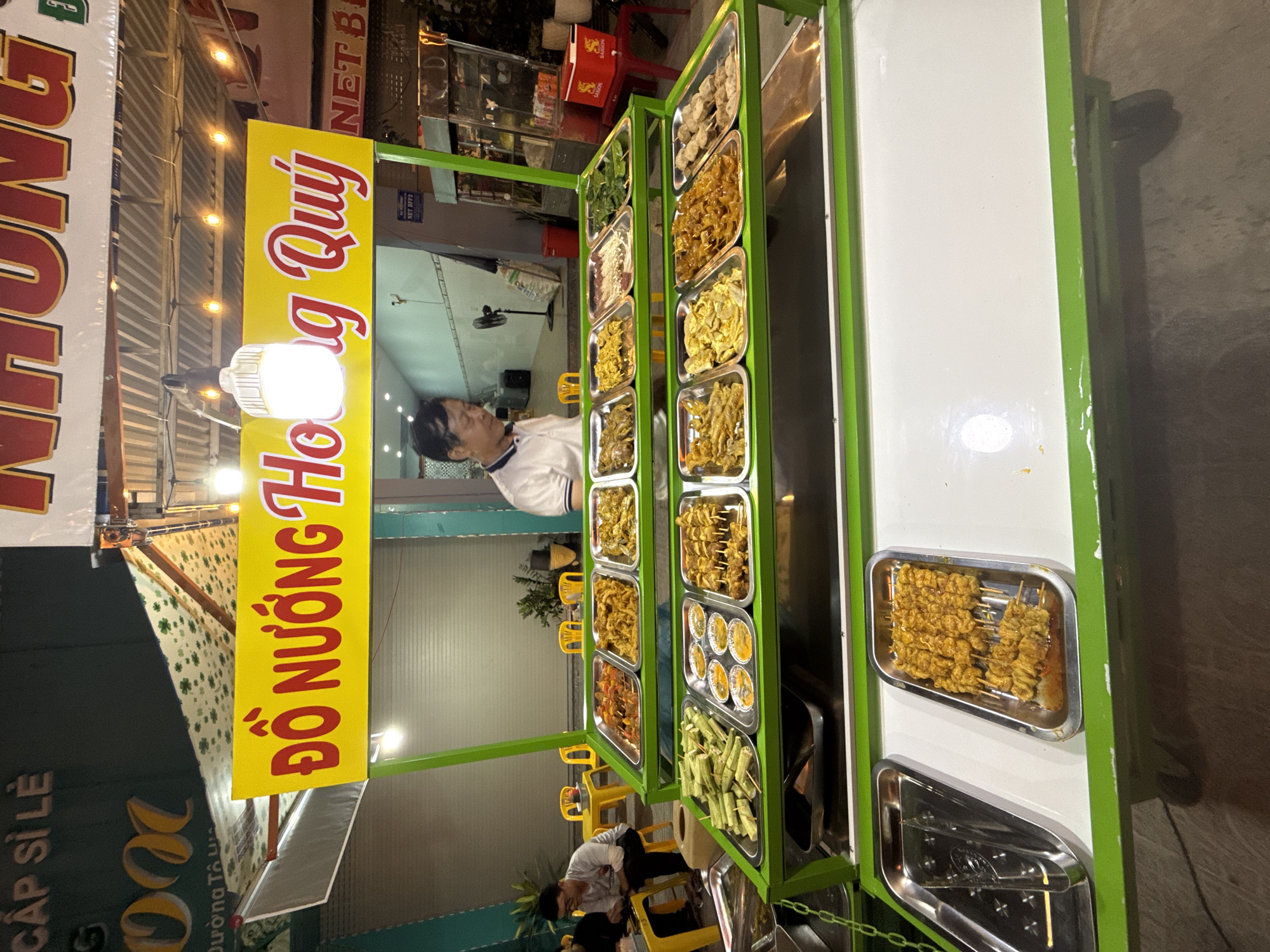The image captures a street food stall in Nha Trang Ward, Vietnam, specializing in grilled items. The stall is brightly lit, featuring a prominent yellow sign with red text that reads "ĐỒ NƯỚNG HOÀNG QUÝ," meaning "Hoàng Quý Grilled Food." This sign is illuminated by a string of small lights and a larger, modern LED light fixture.
The stall is set up with multiple tiers of metal trays displaying various food items, primarily skewers of marinated meat and other grilled ingredients. On the top tier, trays hold what appear to be skewers of chicken or pork, some with vegetables. The middle tier showcases a wider variety, including what looks like rolled spring rolls, small bowls with dipping sauces, and more skewers of meat. The bottom tier is a large, empty stainless steel tray, likely for serving or holding prepared food.
A man, presumably the vendor, is visible behind the stall, wearing a white polo shirt and looking down at the food. He appears to be of Asian descent and has dark hair. In the background, yellow plastic tables and chairs are set up, suggesting a seating area for customers. Other elements of the environment include a wall with a closed metal shutter and some potted plants, typical of a street-side establishment. The overall impression is a bustling, nighttime food stall ready to serve customers.