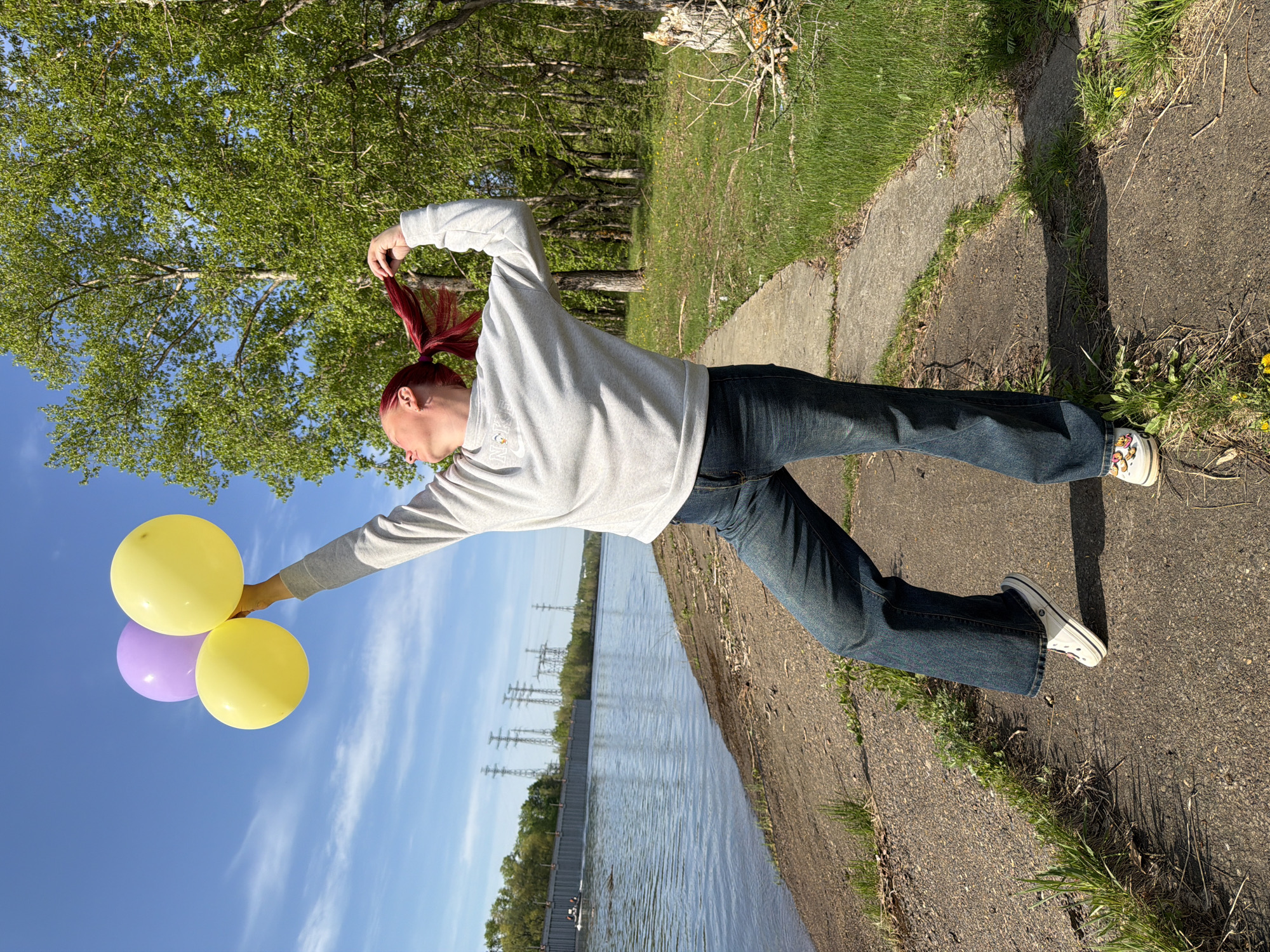 A young person with red hair is standing on a sidewalk in Chaikovsky, Russia, holding balloons. The sidewalk is next to a body of water with power lines in the background. Trees and grass are on the other side of the sidewalk. The person is wearing a gray sweater, jeans, and sneakers.